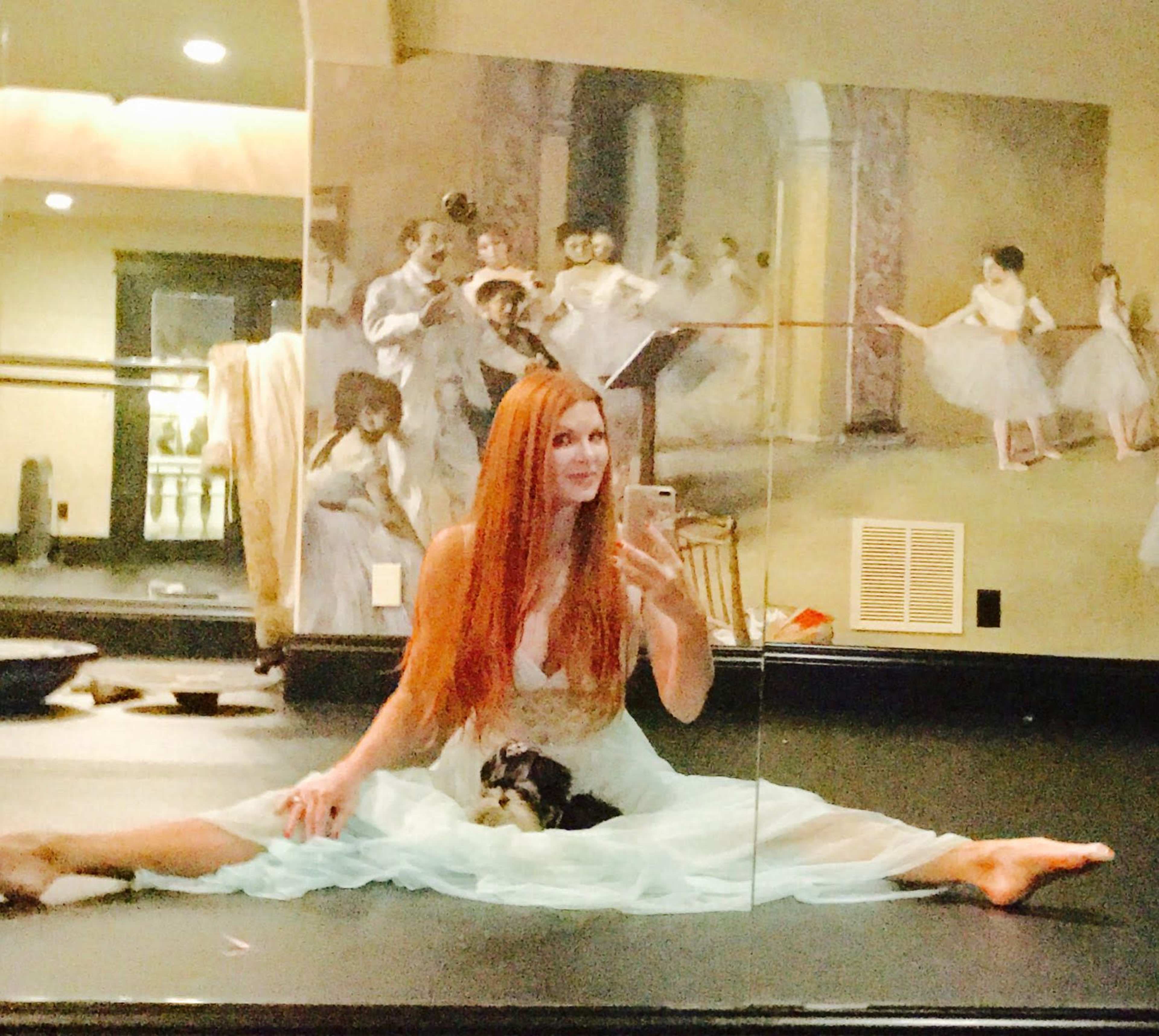 A woman with long red hair sits in a split on a dance studio floor, holding a small dog, with a mirror reflecting a painting of ballet dancers in the background.