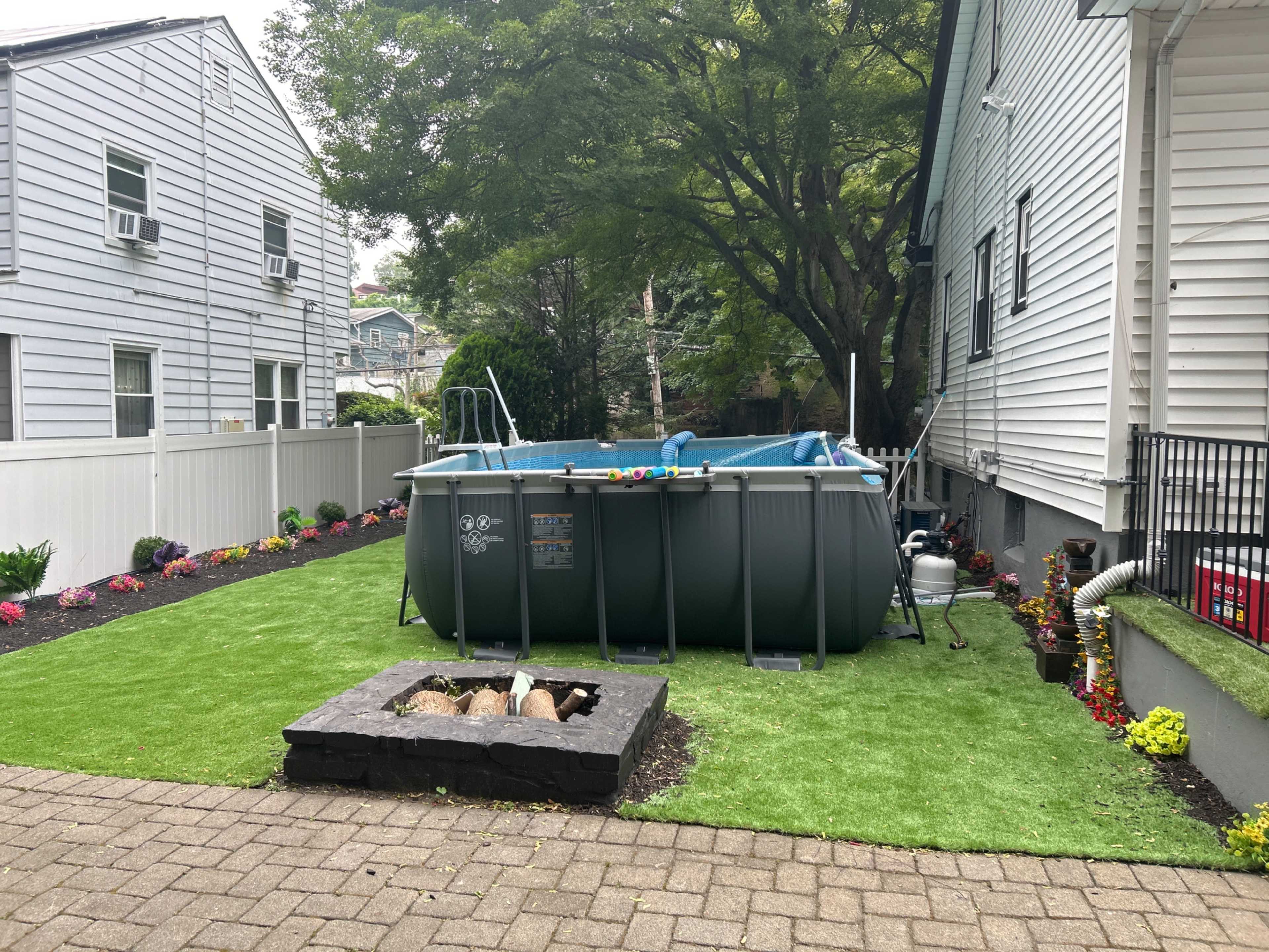 A backyard features an above-ground pool surrounded by green turf and a fire pit, with floral landscaping and a neighboring fence.