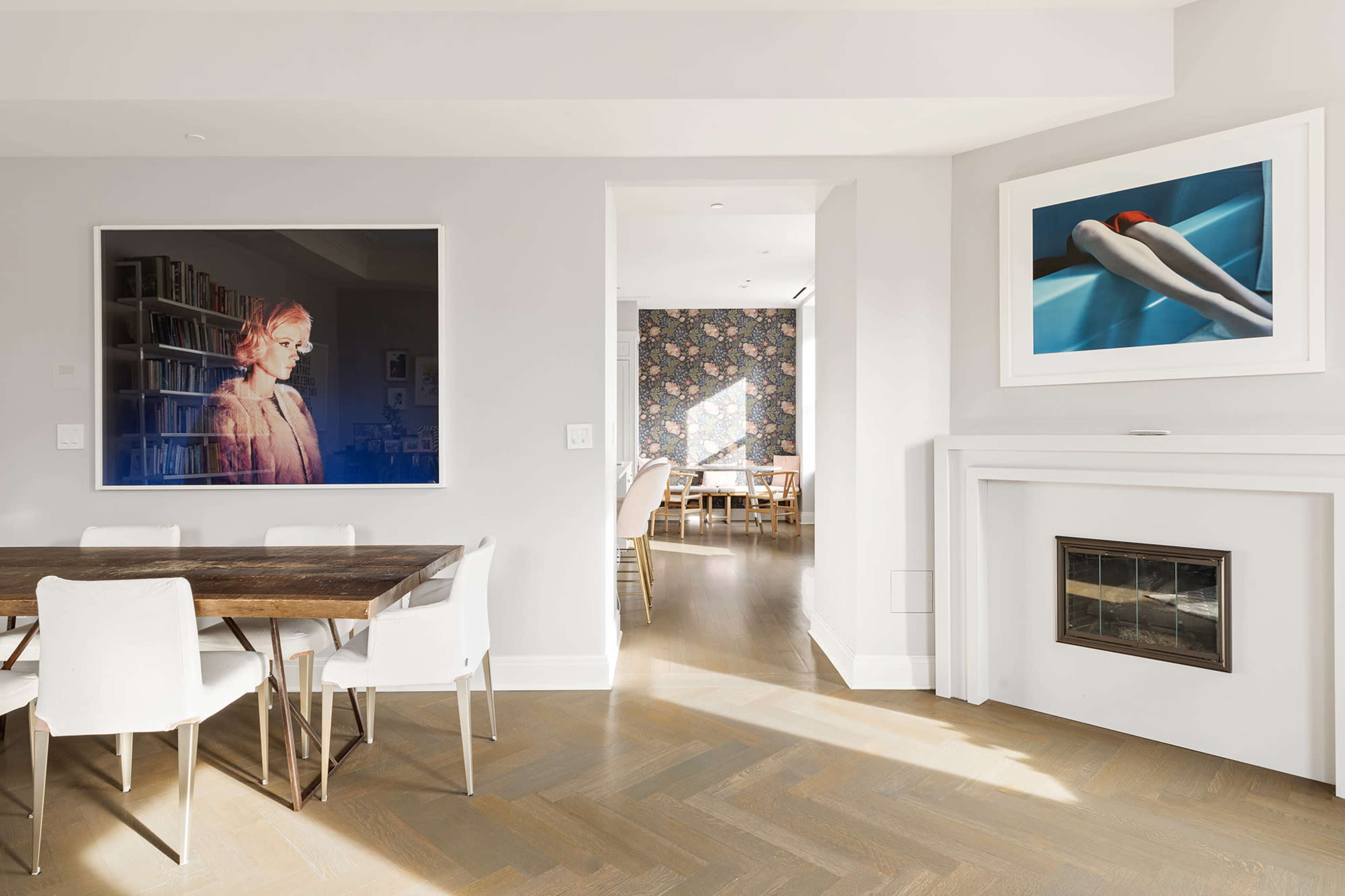 The image shows a modern interior space featuring a dining area with a wooden table and white chairs, framed photographs on the walls, and a glimpse of a floral wallpapered room through an open doorway.