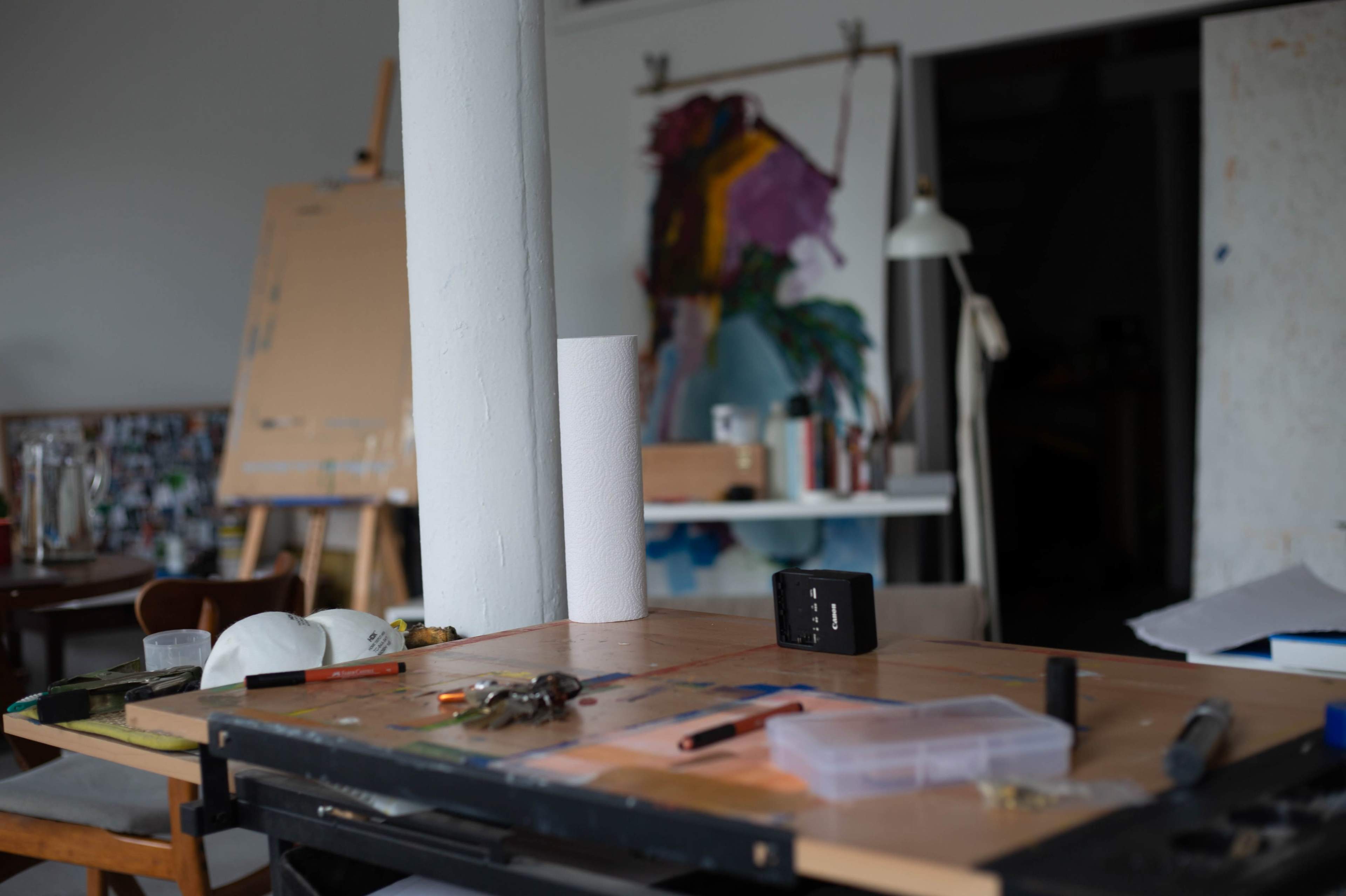 The image shows an artist's studio with a cluttered worktable, featuring various tools, supplies, and a partially visible large canvas in the background.