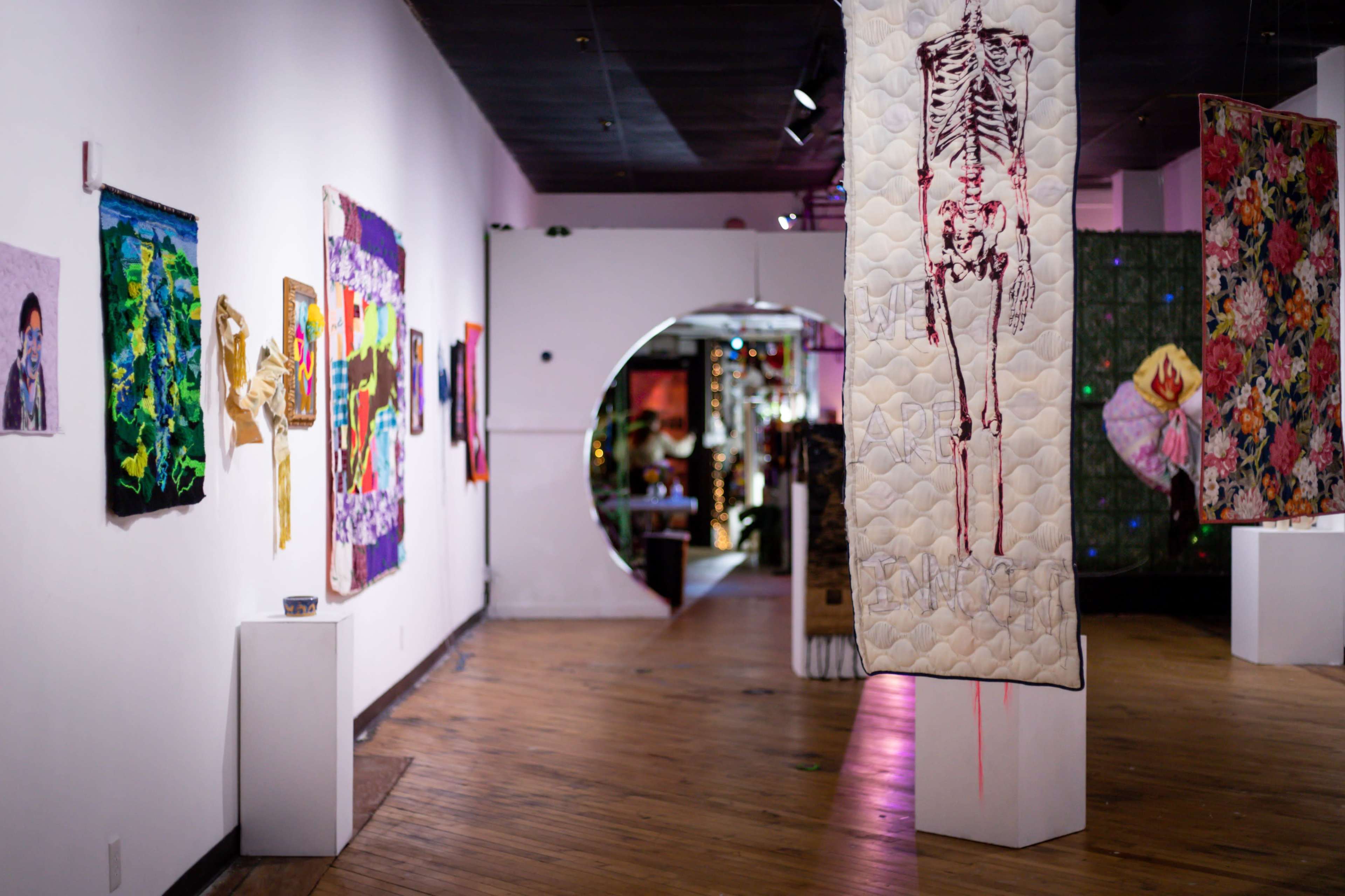 An art gallery featuring various colorful artworks displayed on the walls, with a prominent quilted piece hanging in the center.