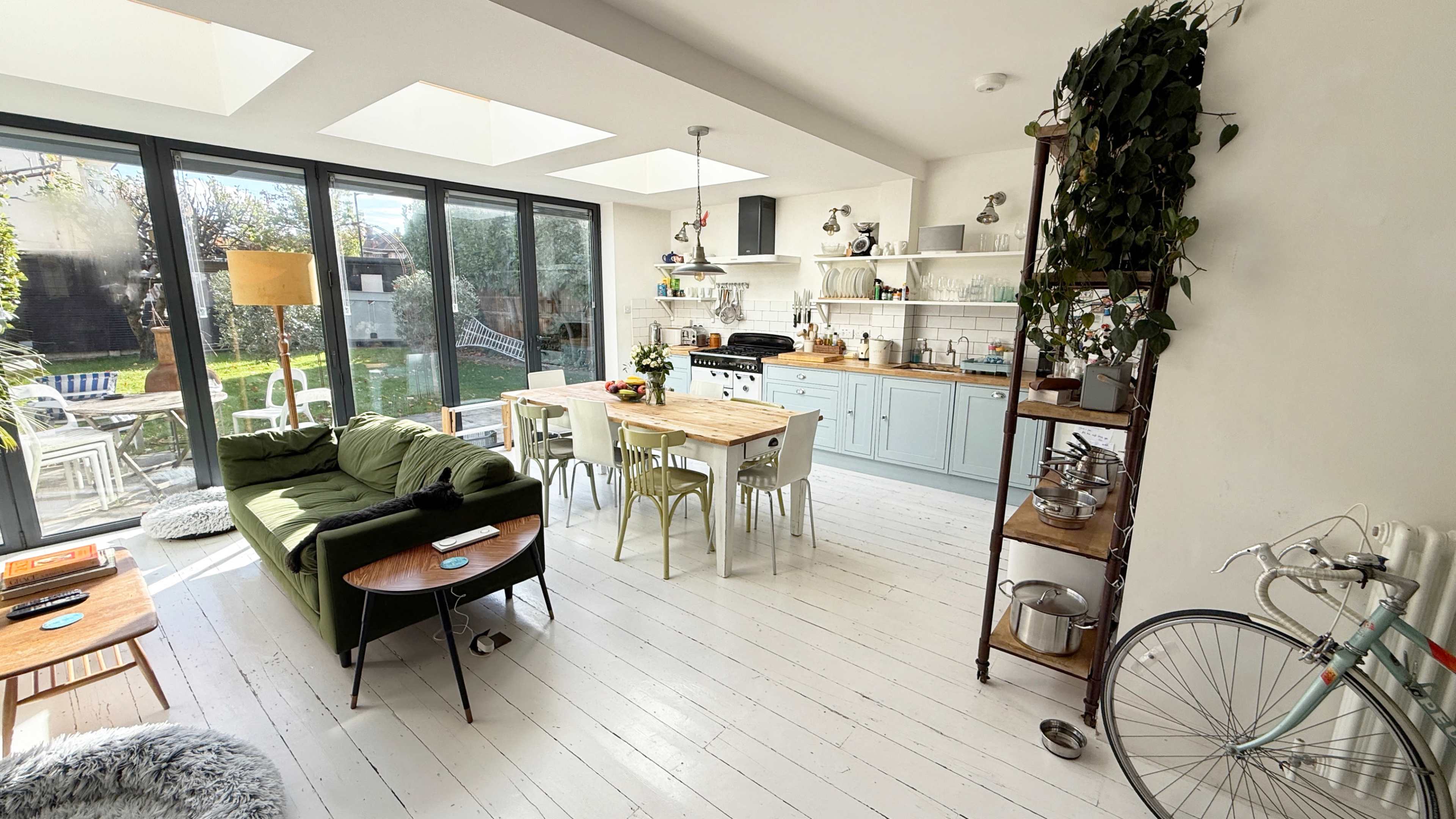 The image shows a modern, open-concept kitchen and dining area with large windows, light wood flooring, and a mixed arrangement of furniture including a green sofa, wooden dining table, and various plants.