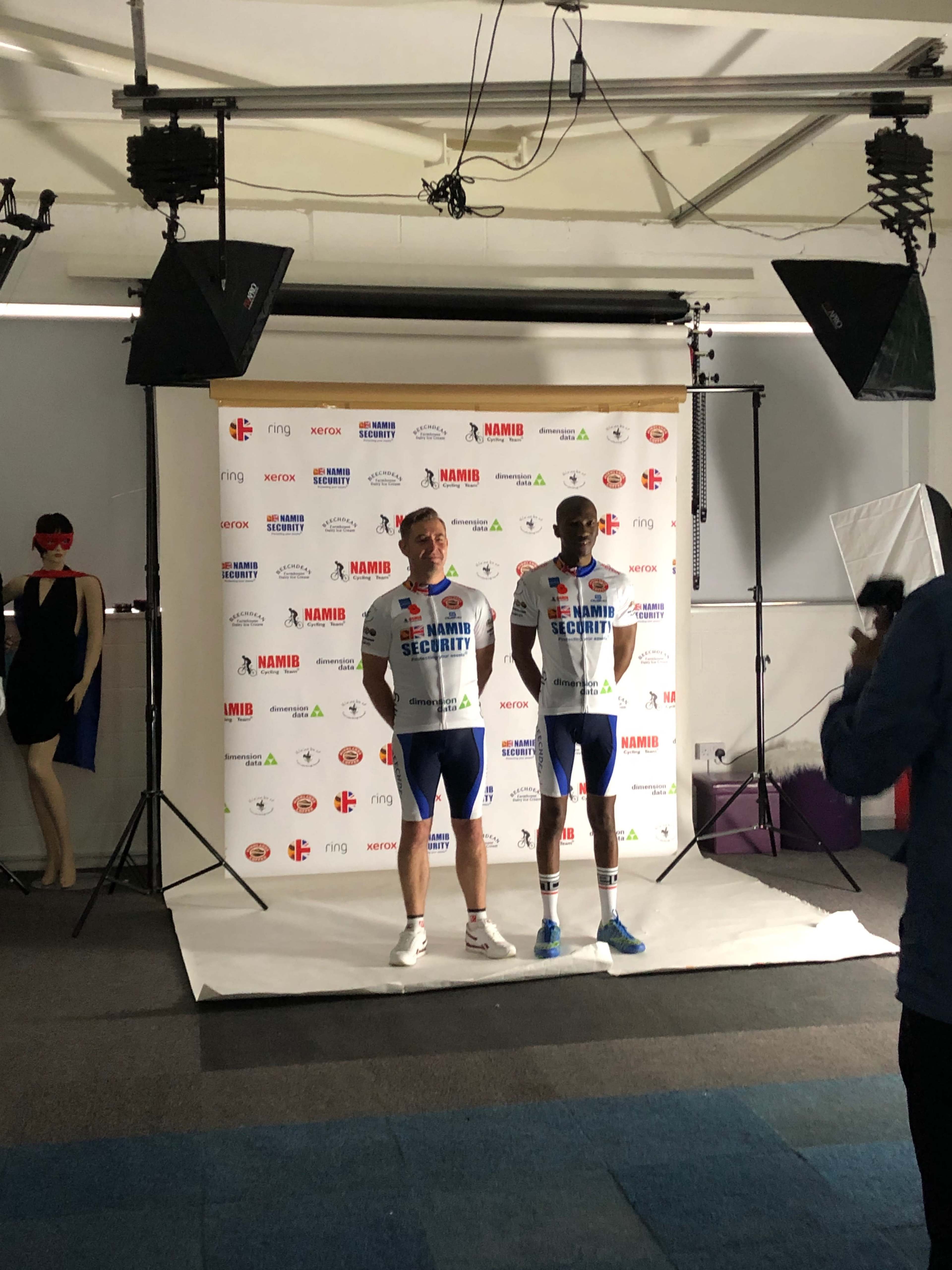 Two athletes pose for a photo in cycling attire in front of a branded backdrop, with a mannequin and lighting equipment visible in the scene.