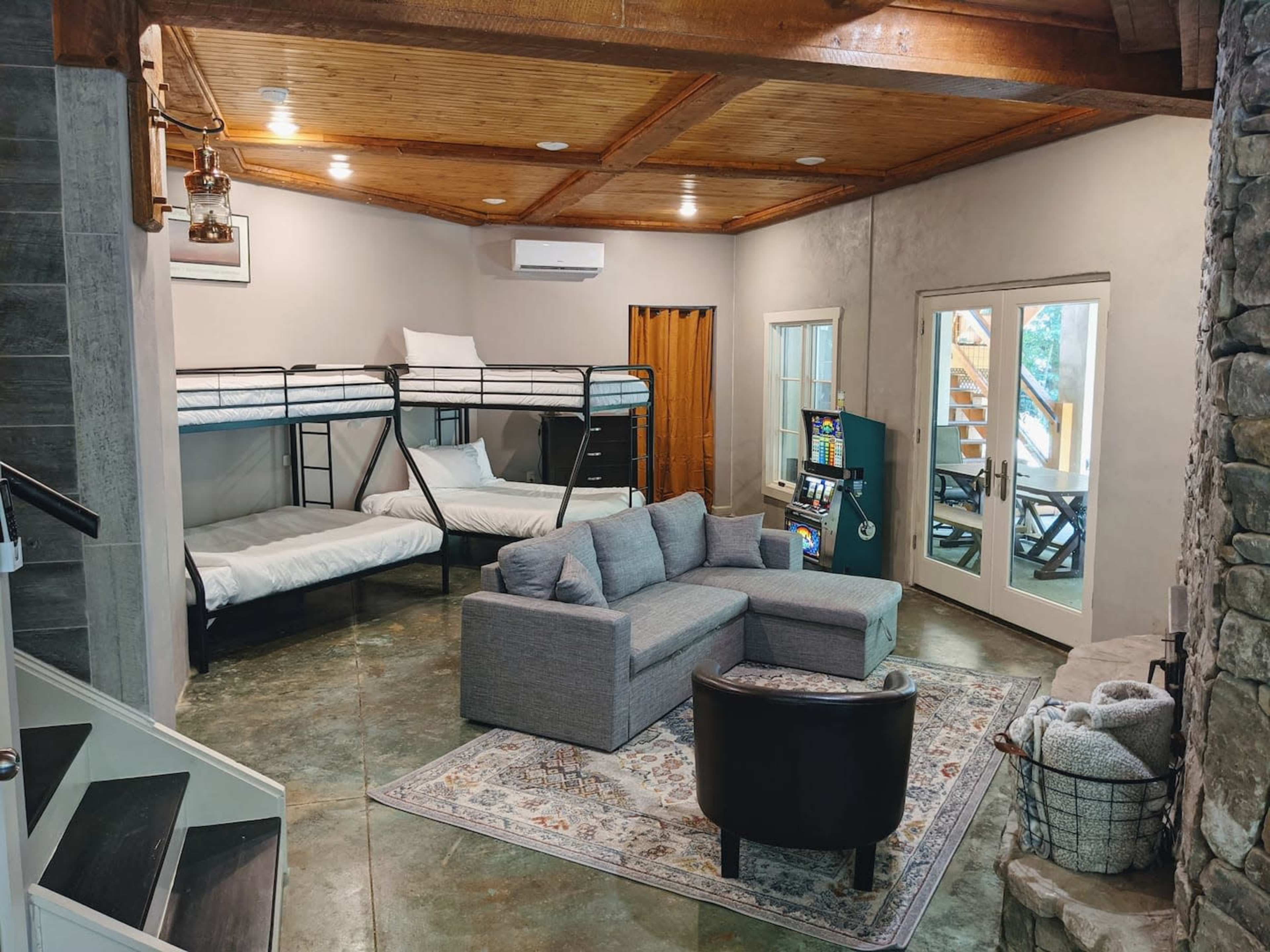 A modern living space featuring a gray sofa, a black armchair, two bunk beds, and a stone fireplace, with large windows and a door leading outside.
