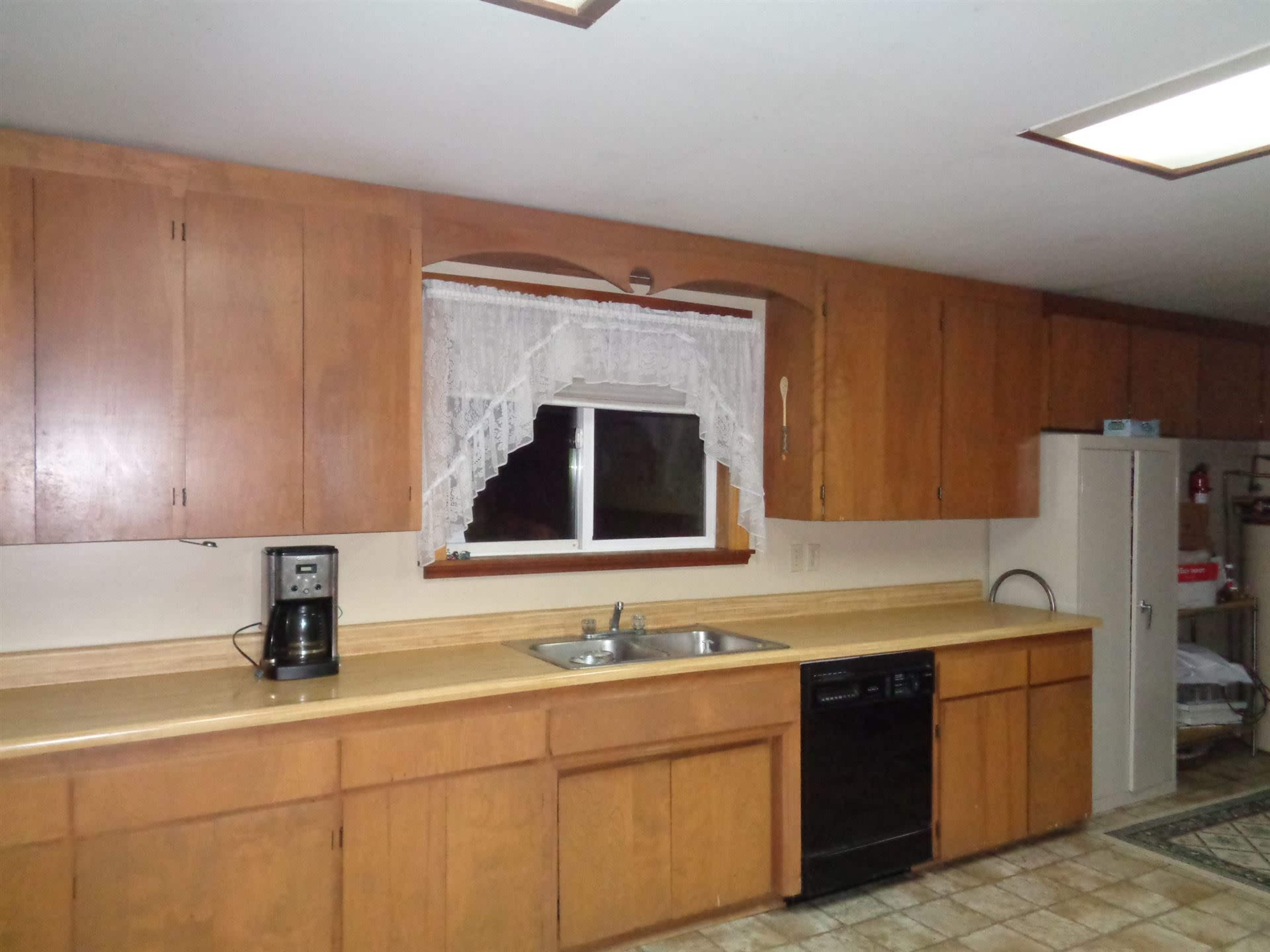 The kitchen features wooden cabinets, a window with lace curtains, a double sink, a coffee maker, a dishwasher, and a refrigerator along the counters.