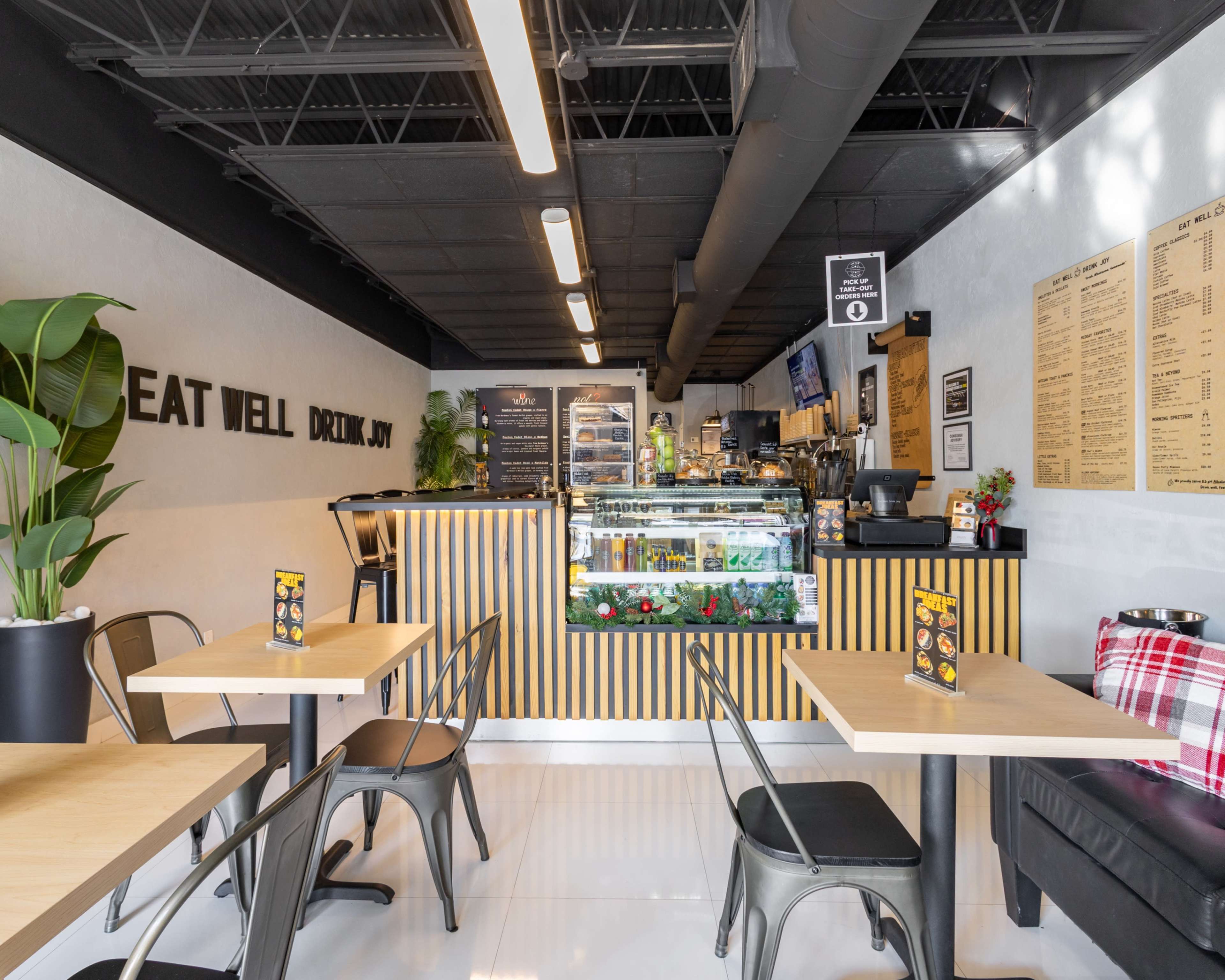 The image shows a modern café with wooden tables, black chairs, and a service counter displaying beverages and snacks.