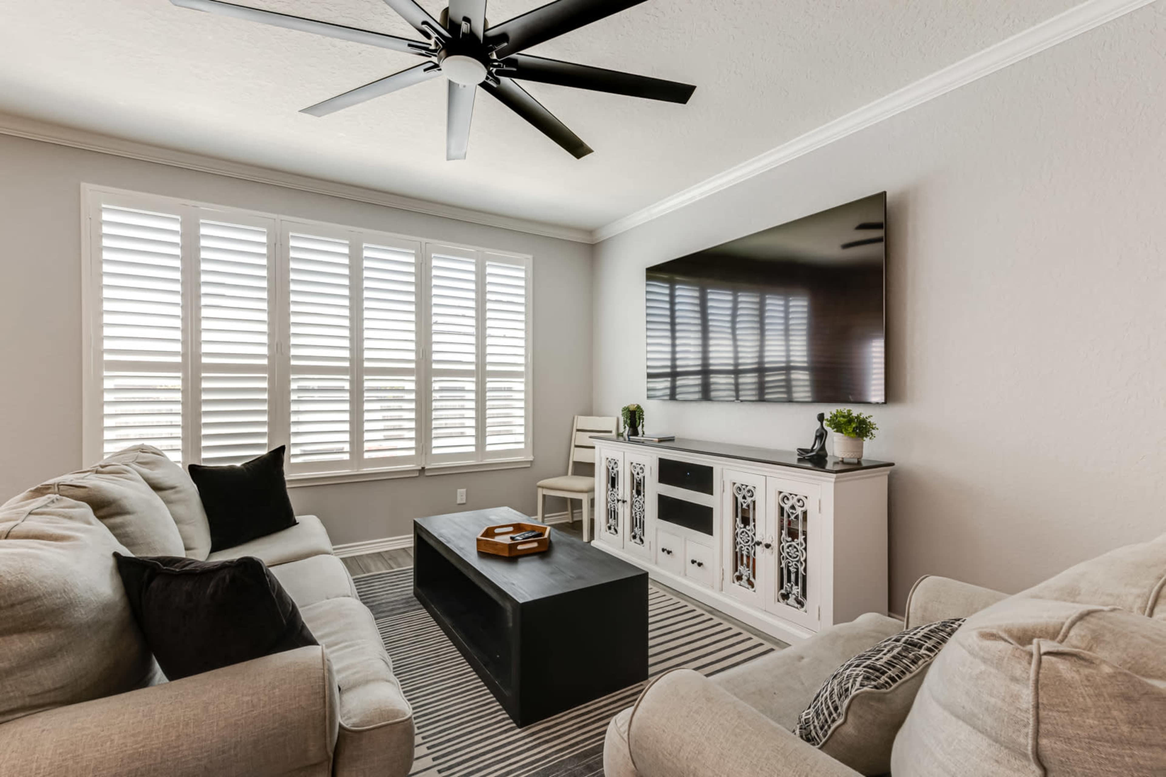 A living room features two sofas, a coffee table, a television mounted on the wall, and shuttered windows.