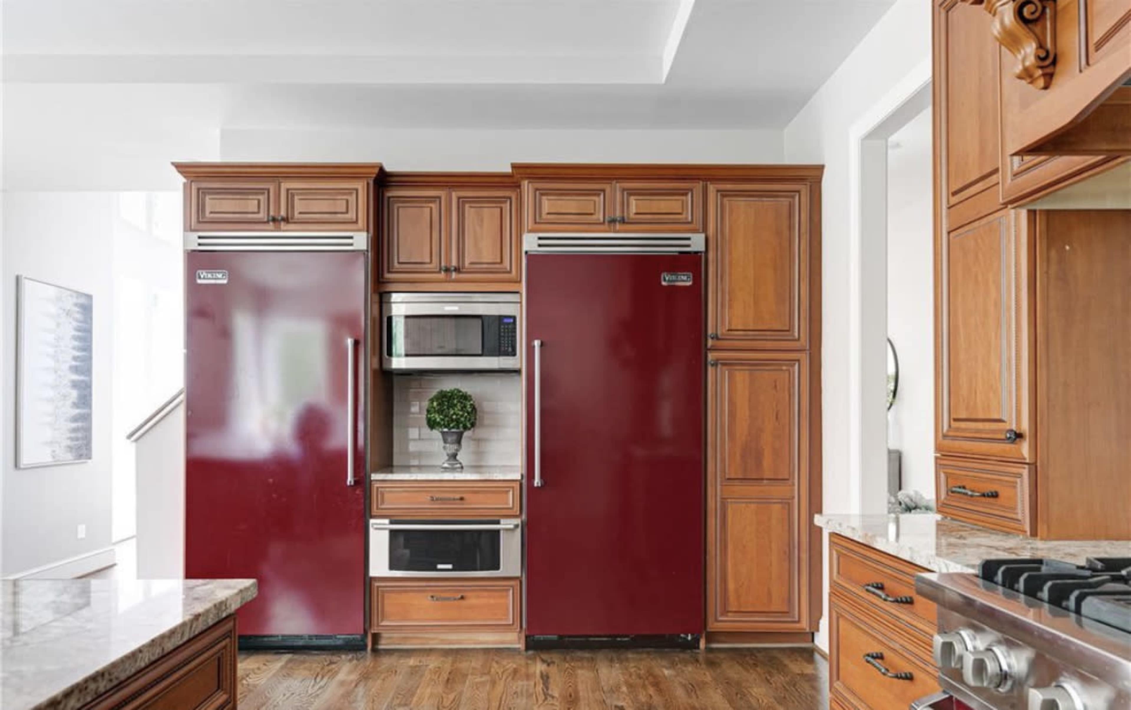 A kitchen with two red refrigerators flanking a built-in microwave and a small plant on a countertop beneath wooden cabinetry.