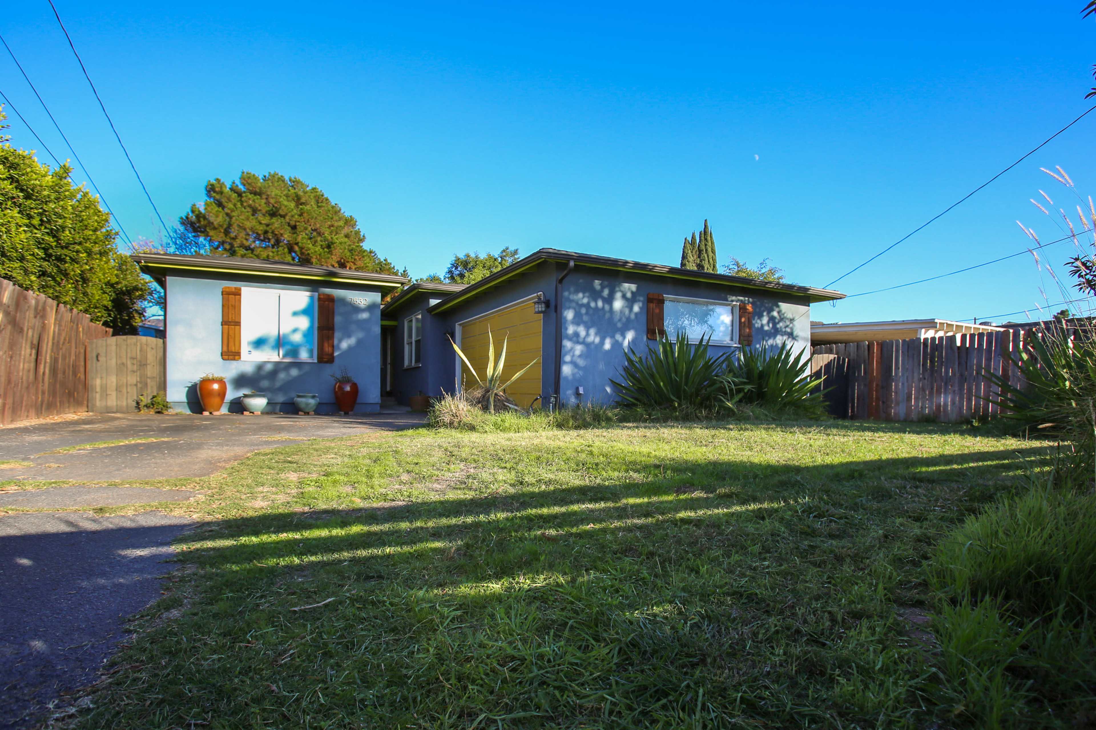 A blue house with yellow accents sits at the end of a grassy driveway, surrounded by low shrubs and a wooden fence.