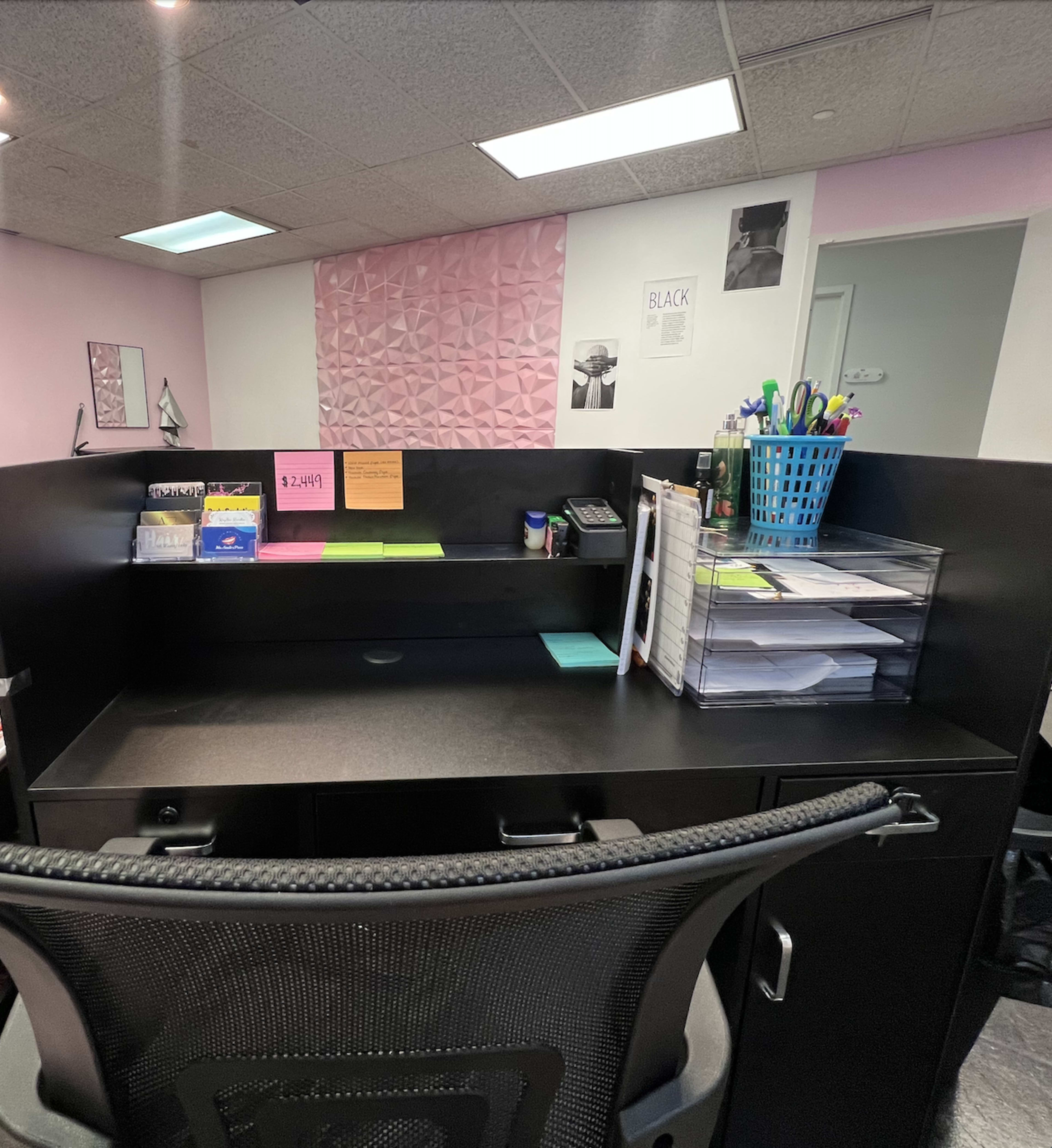 A black office desk with a computer chair, organized papers, a blue basket, and a pink textured wall in the background.