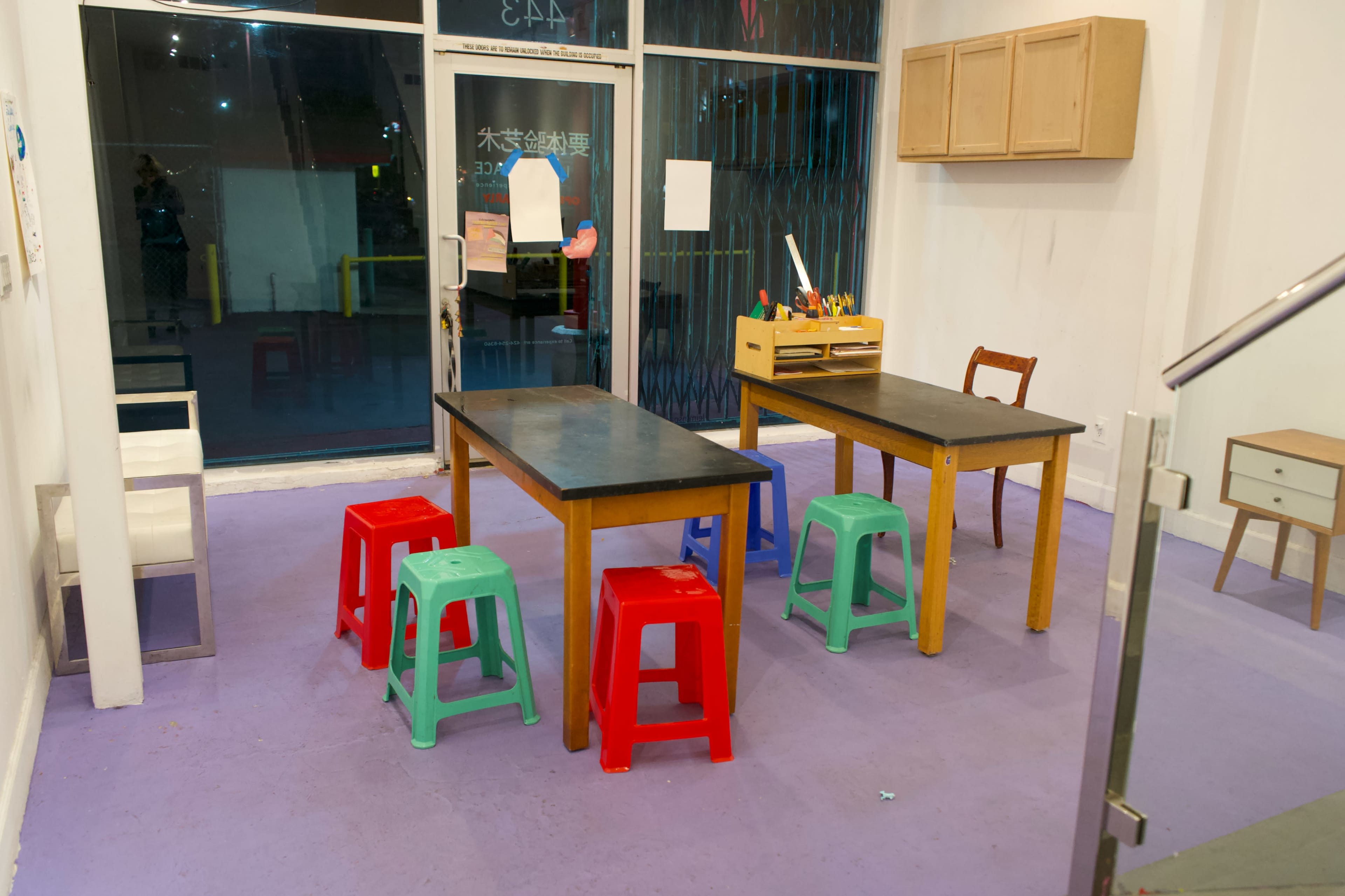 The scene shows a small room with two tables surrounded by colorful plastic stools and a view of a large window.