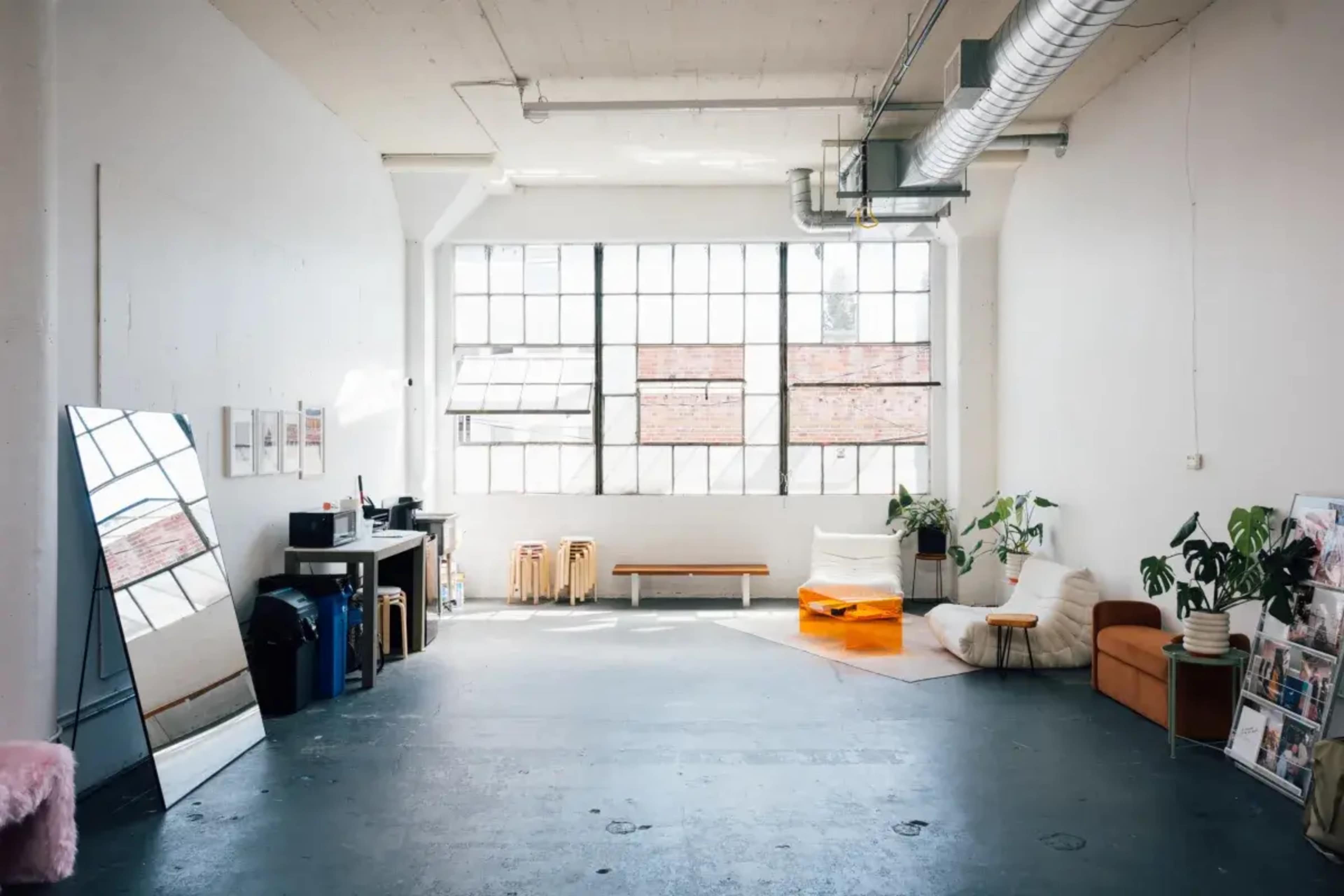 The image shows a spacious, minimalist room featuring large windows, a few pieces of furniture including a sofa and a stool, and a mirror against one wall.
