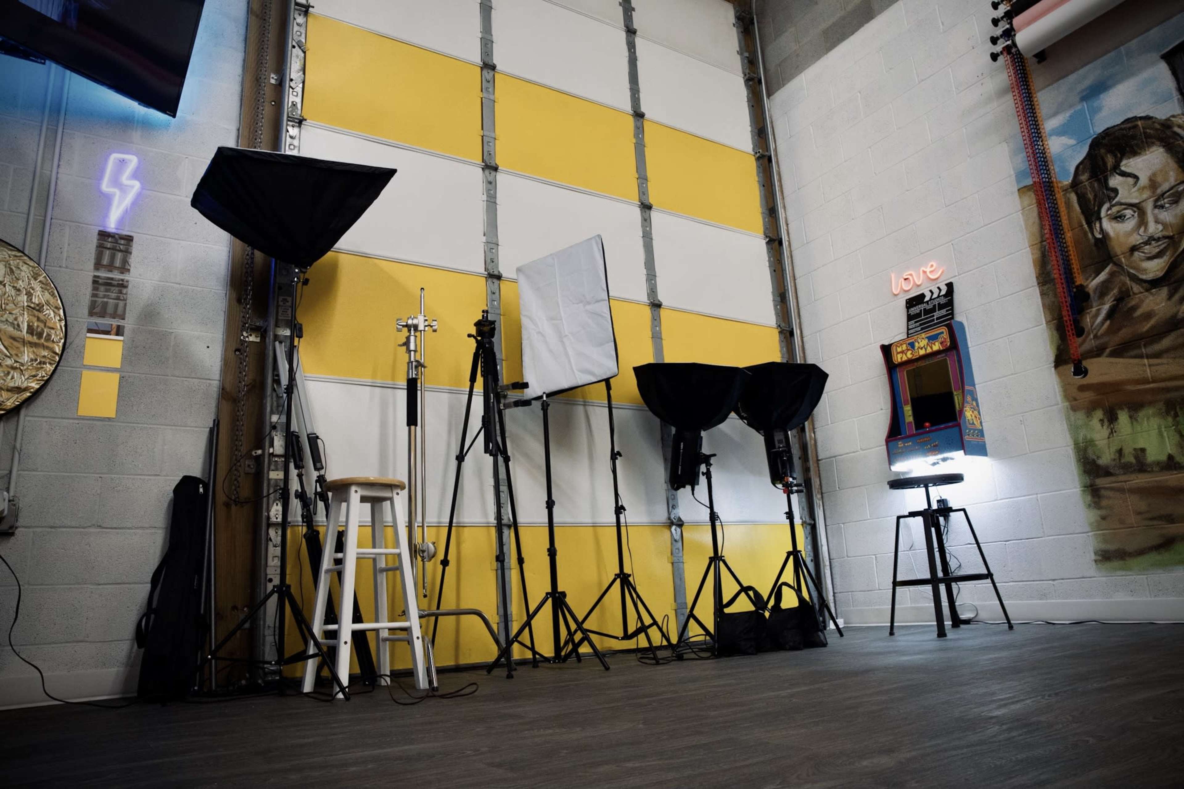 A photography studio with black lighting equipment, a tall stool, and an arcade game machine against a backdrop of yellow and gray walls.