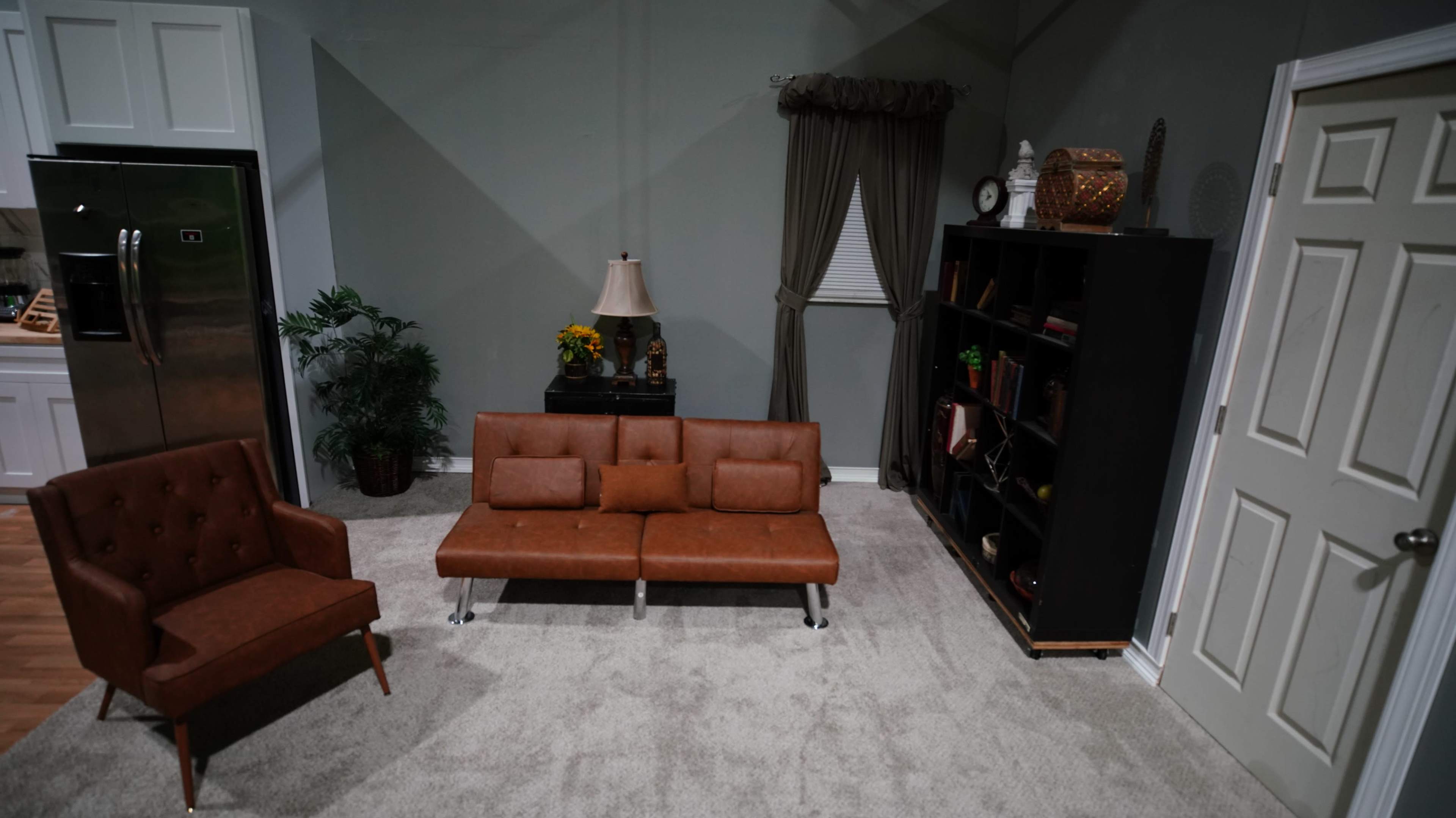 A modern living room features a brown leather sofa and armchair, a bookshelf filled with items, and a curtain beside a window.