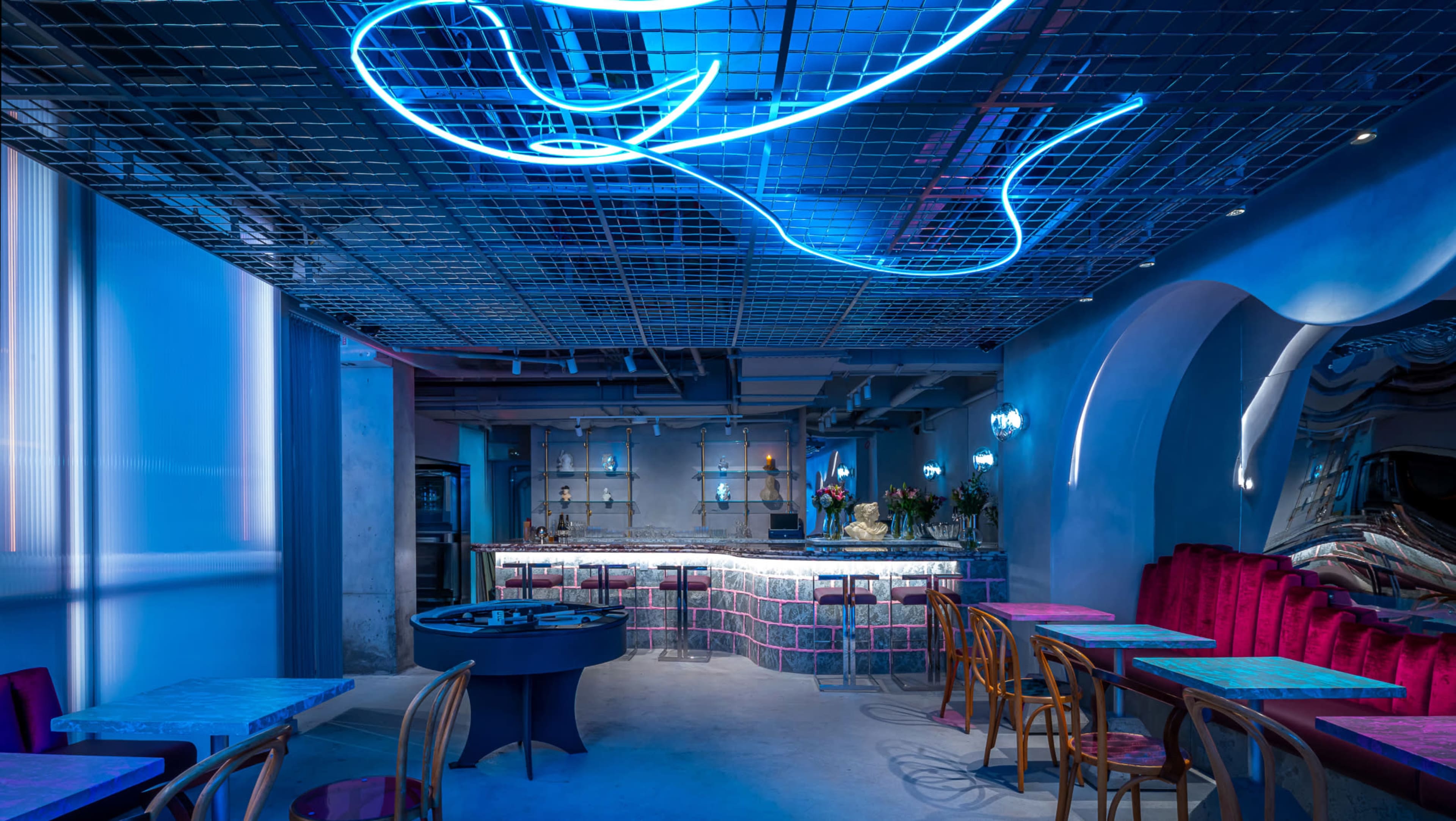 The image shows a modern restaurant interior with blue neon lighting, a grid ceiling, and tables arranged throughout the space.