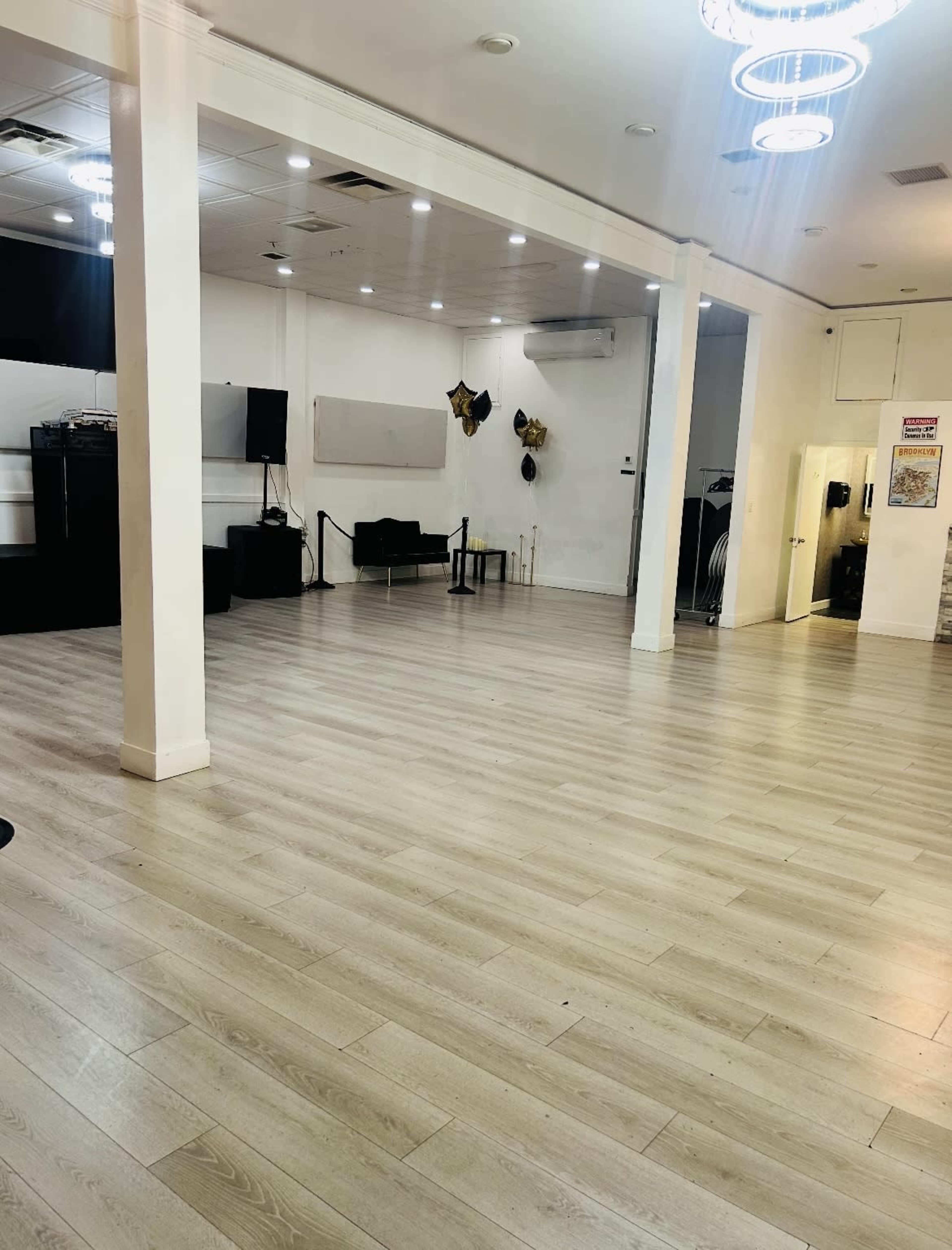 The image shows an empty indoor space with light wooden flooring, white walls, and several lighting fixtures hanging from the ceiling.