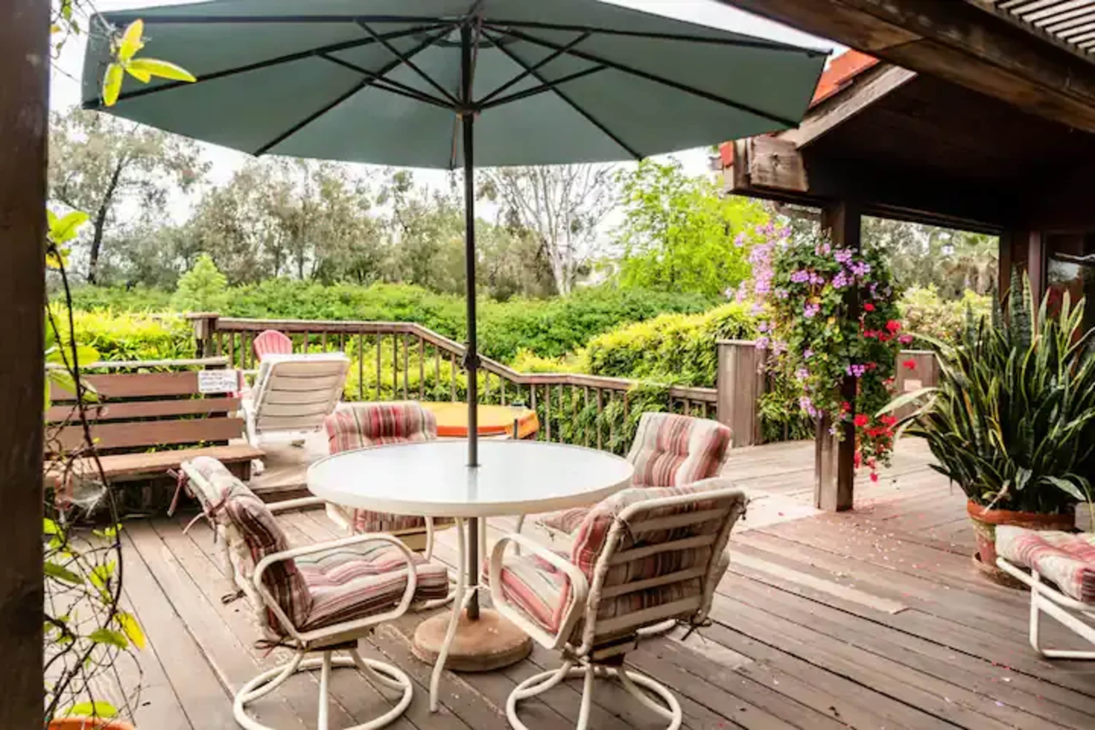 A patio area features a round table with chairs beneath a large umbrella, surrounded by greenery and flowering plants.
