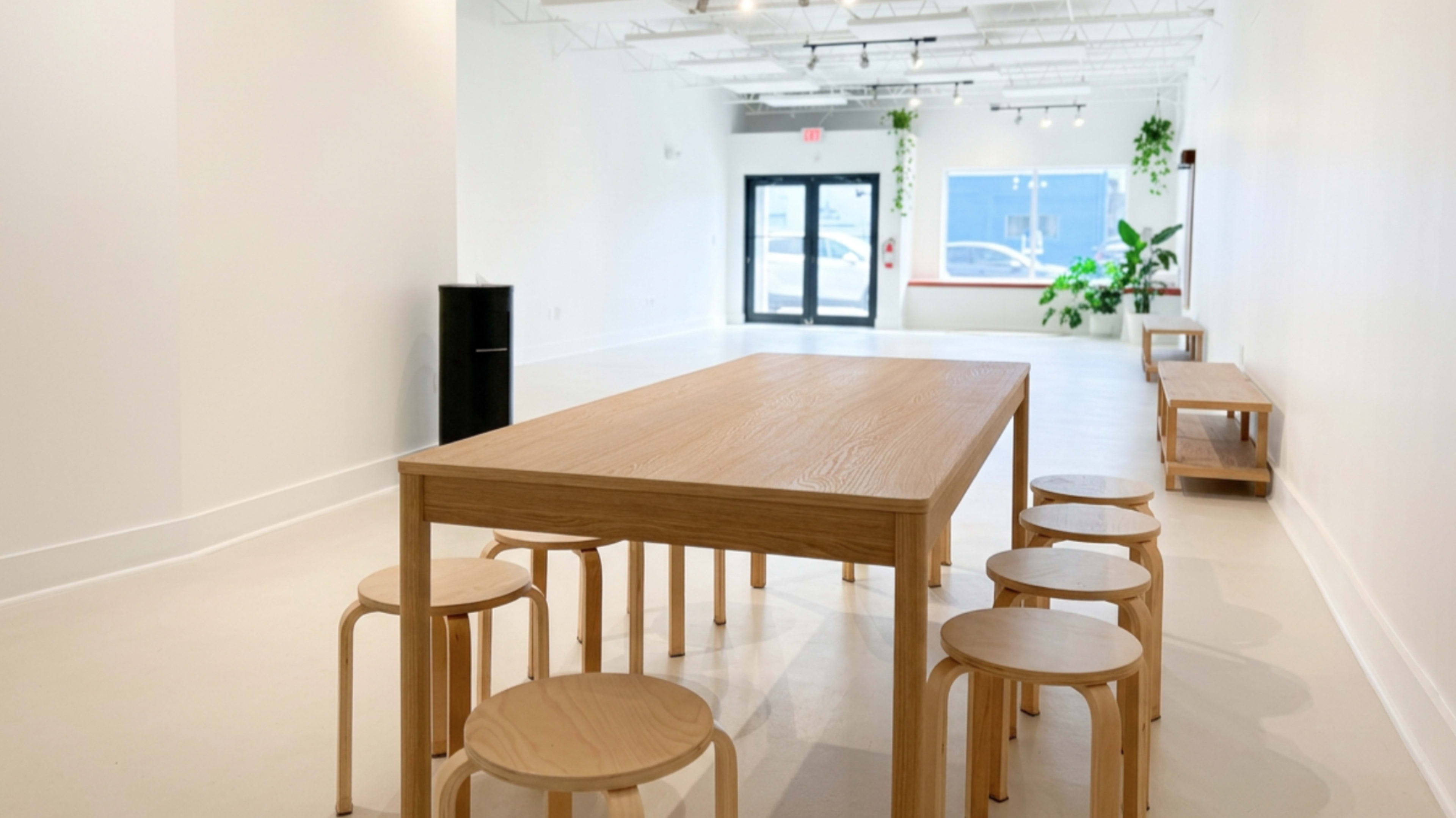 The image shows a minimalist interior space featuring a large wooden table surrounded by several wooden stools, with large windows allowing natural light to enter.