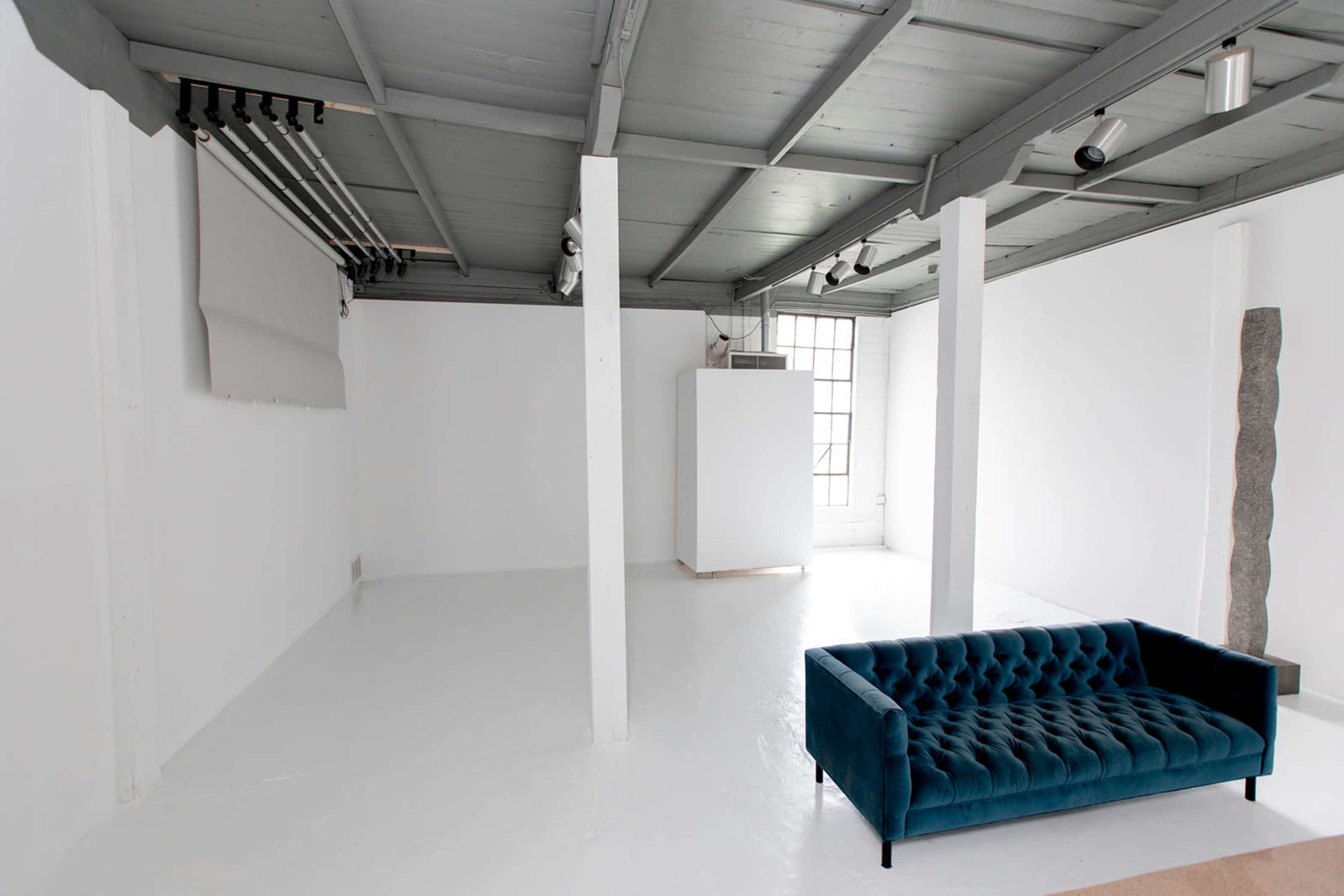 The image shows a minimalist interior space with white walls, a light-colored floor, and a blue tufted sofa in the foreground.