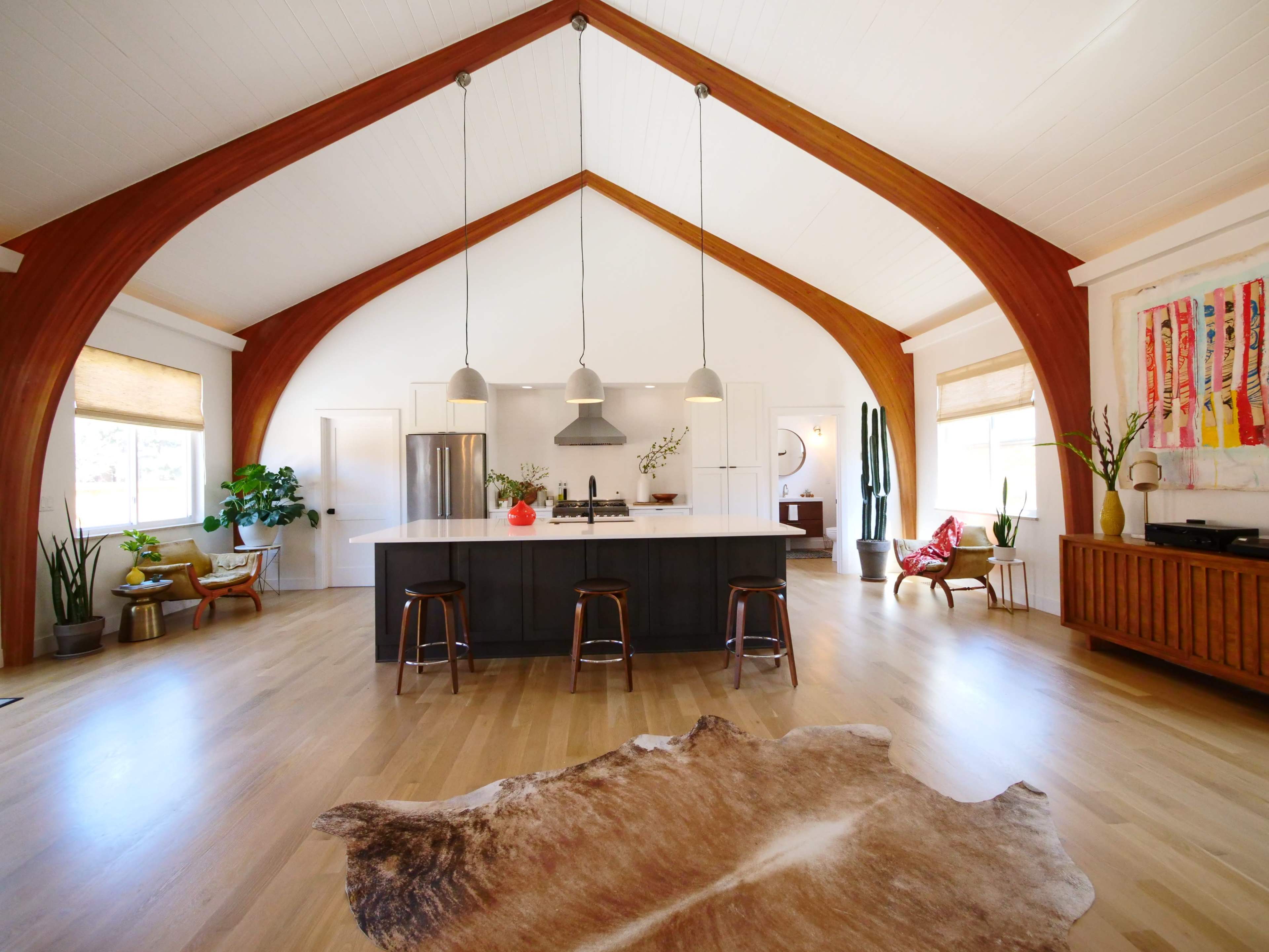 The spacious interior features a large kitchen island with seating, high arched wooden beams, and large windows that illuminate the room.