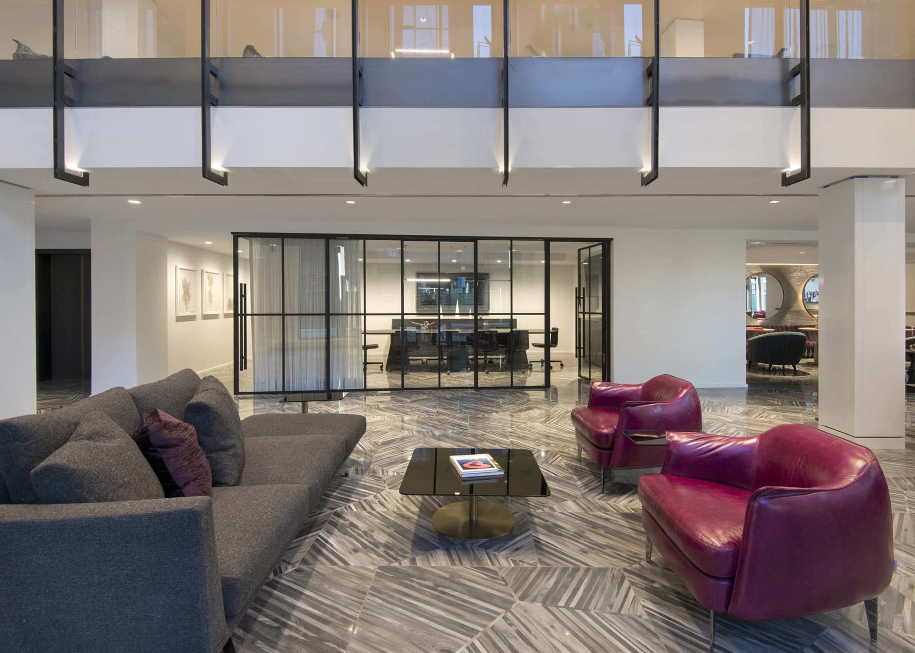 A modern seating area features a dark gray sofa and two purple chairs arranged around a circular coffee table, with a glass-enclosed meeting room visible in the background.