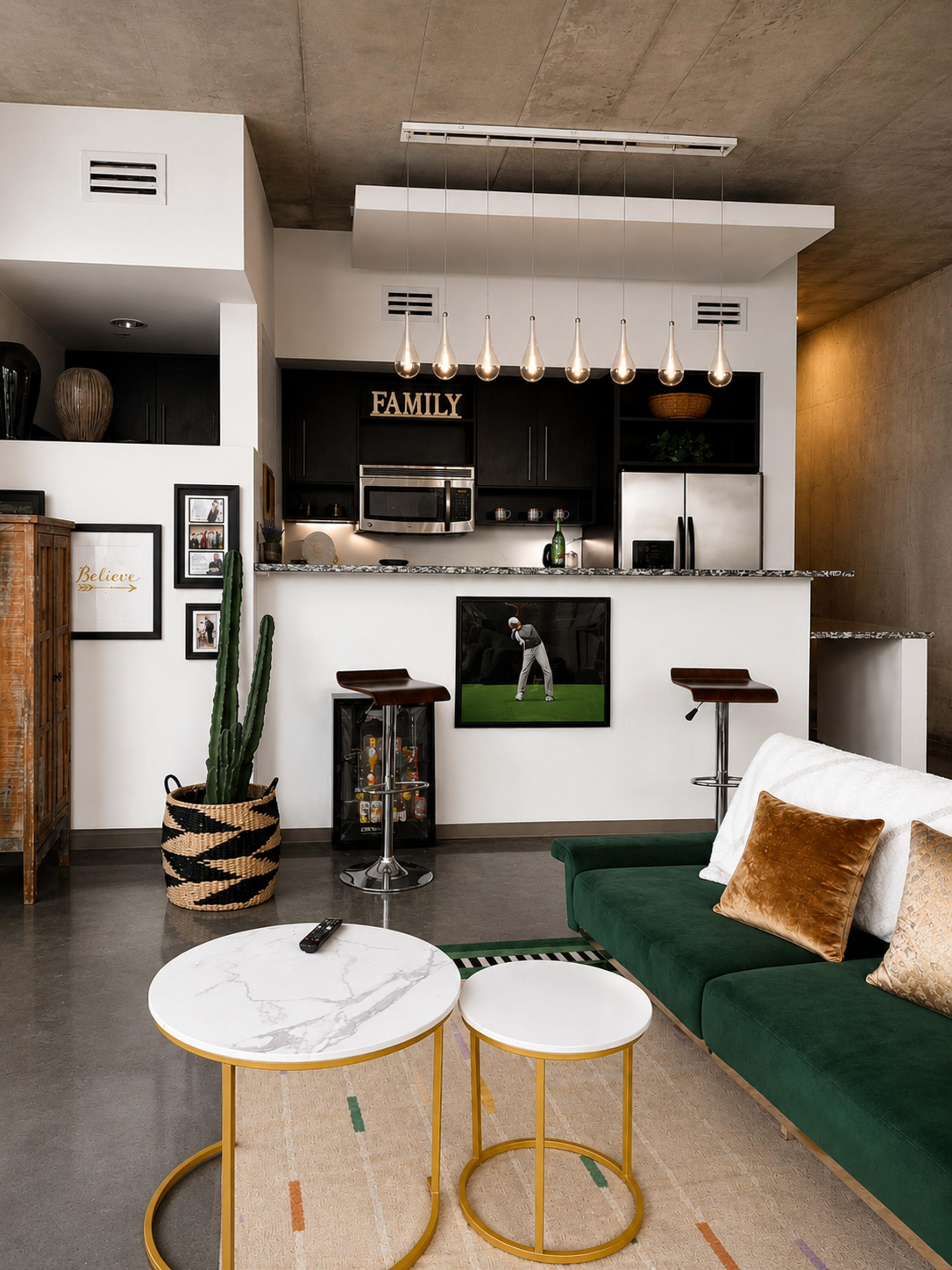 The image shows a modern living room with a green sofa, two round coffee tables, and a kitchen in the background featuring black cabinetry and pendant lighting.