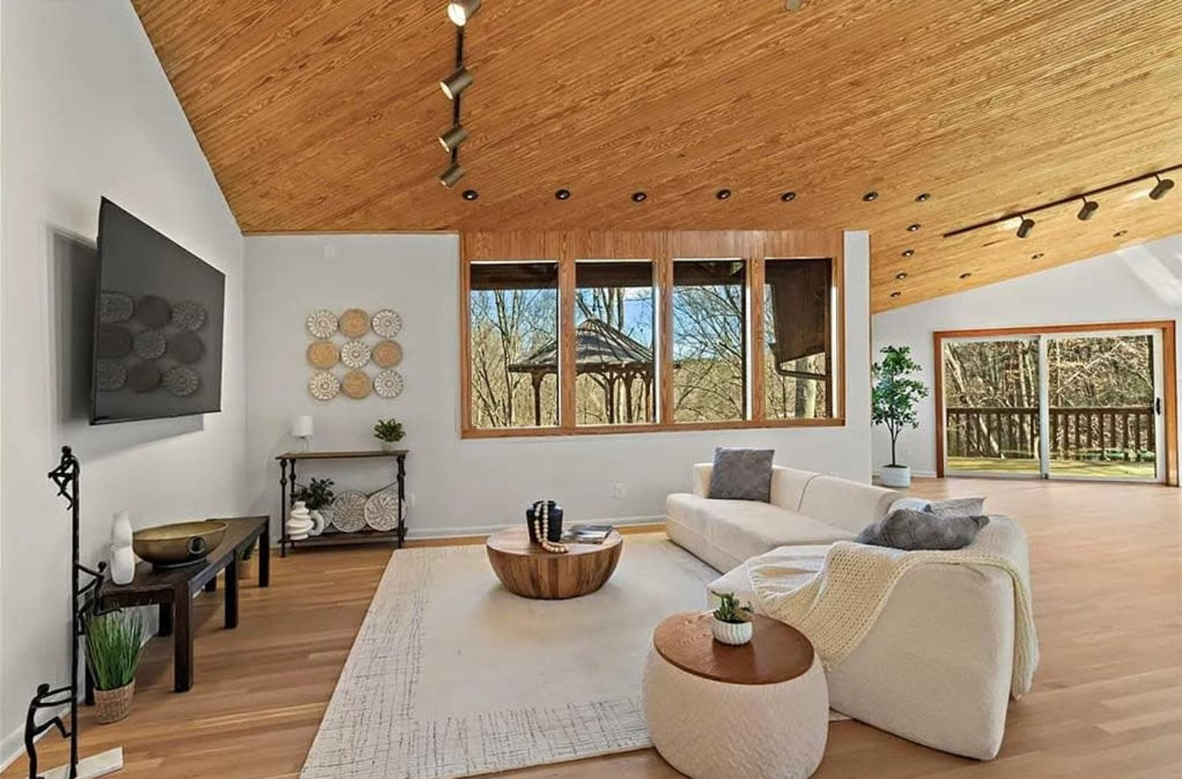 The image shows a modern living room with a high wooden ceiling, large windows overlooking a wooded area, and minimalist furniture.