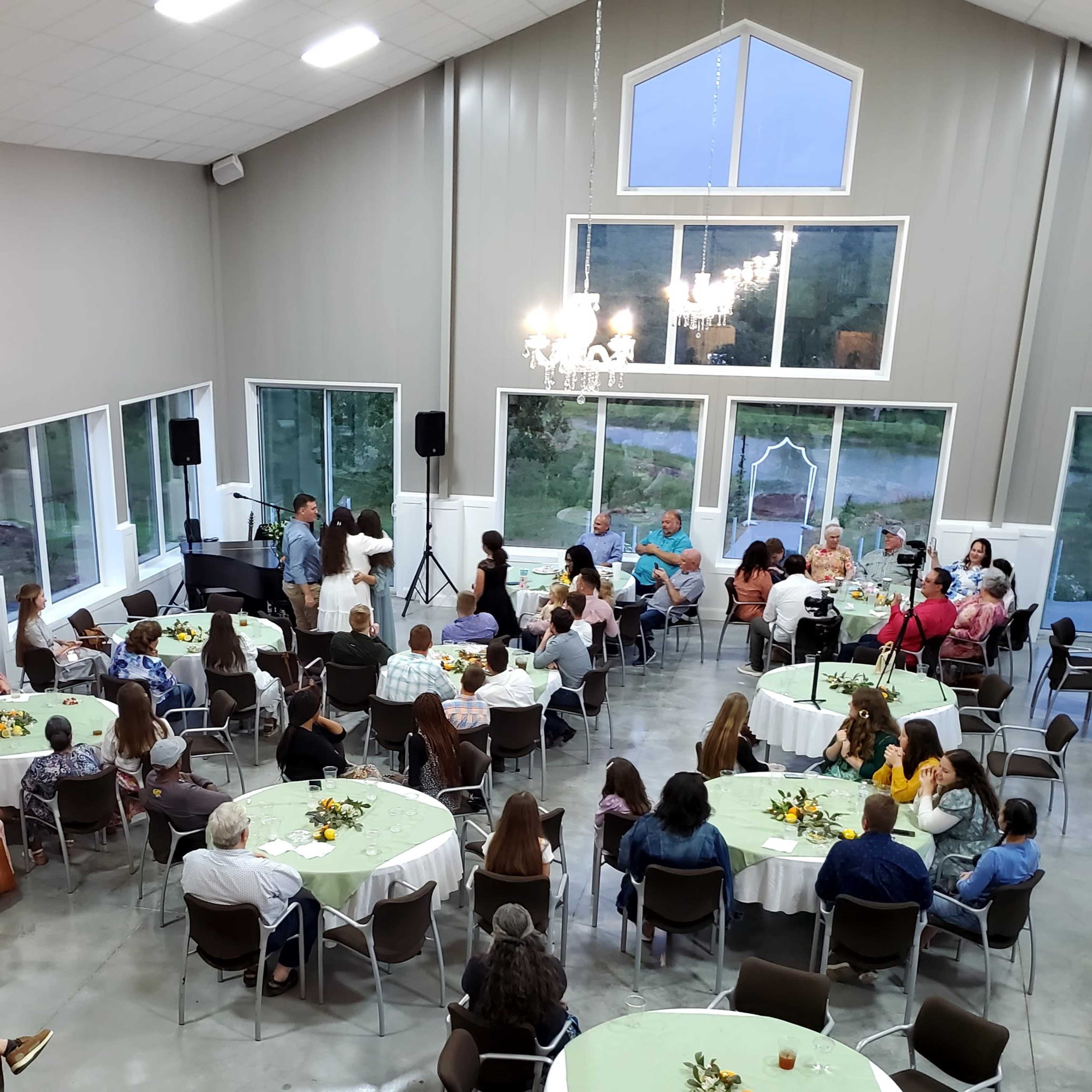 A large gathering takes place in a spacious hall with several round tables arranged for guests and a small stage area for presentations.