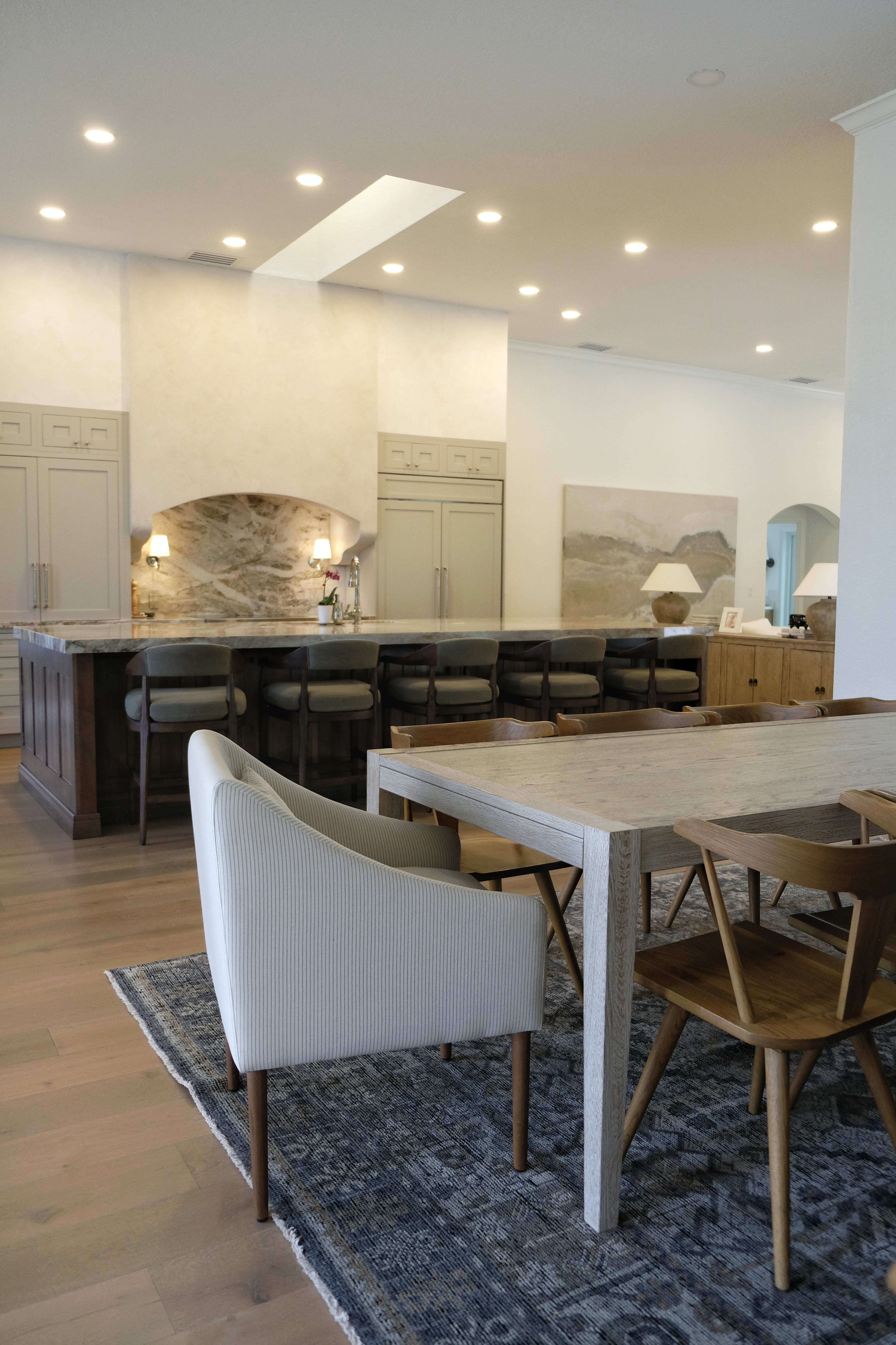 The image shows a modern kitchen and dining area with a large island, dining table, and surrounding chairs on hardwood flooring.