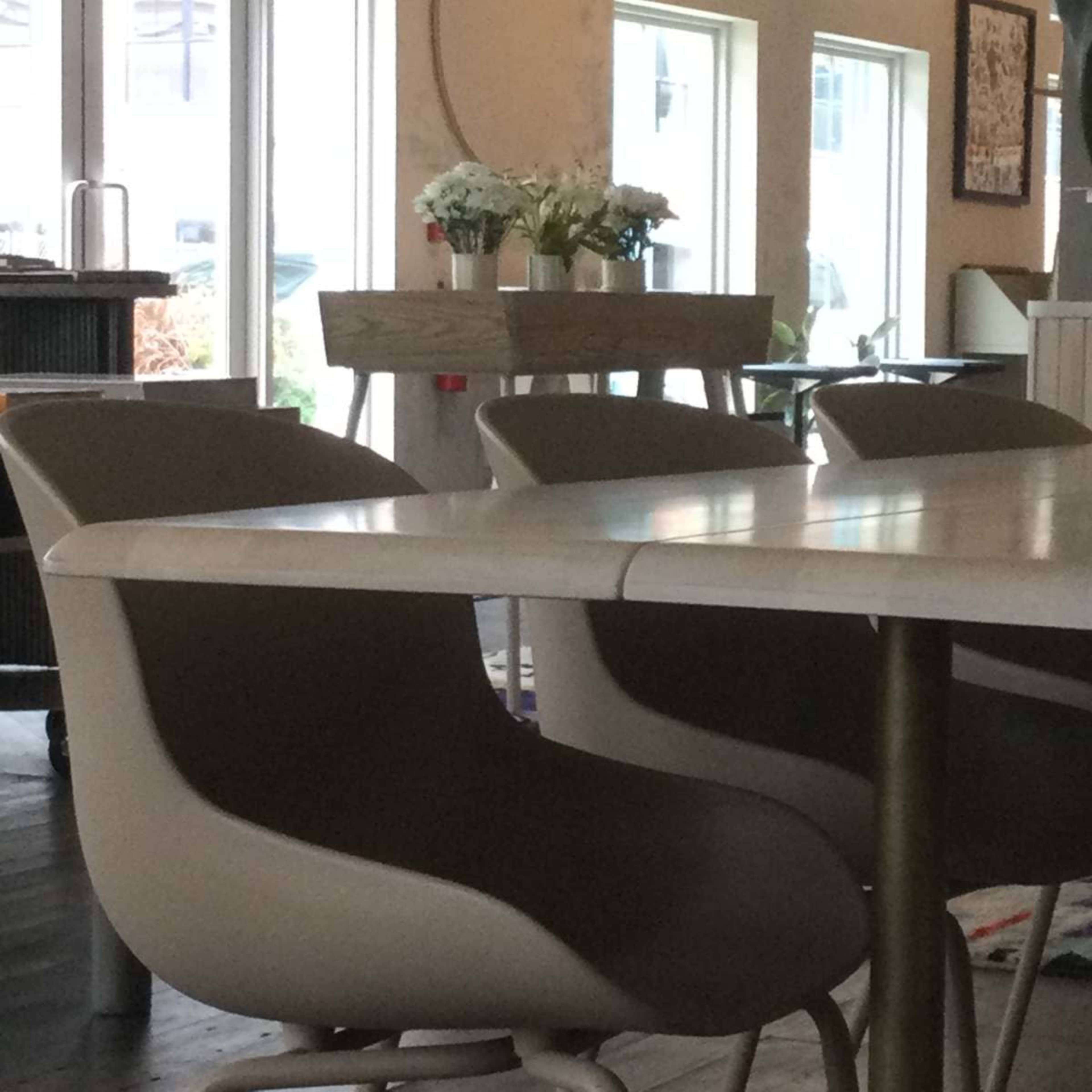 The image features a dining area with a large table and several modern chairs, alongside windows and a decorative arrangement of flowers on a shelf.