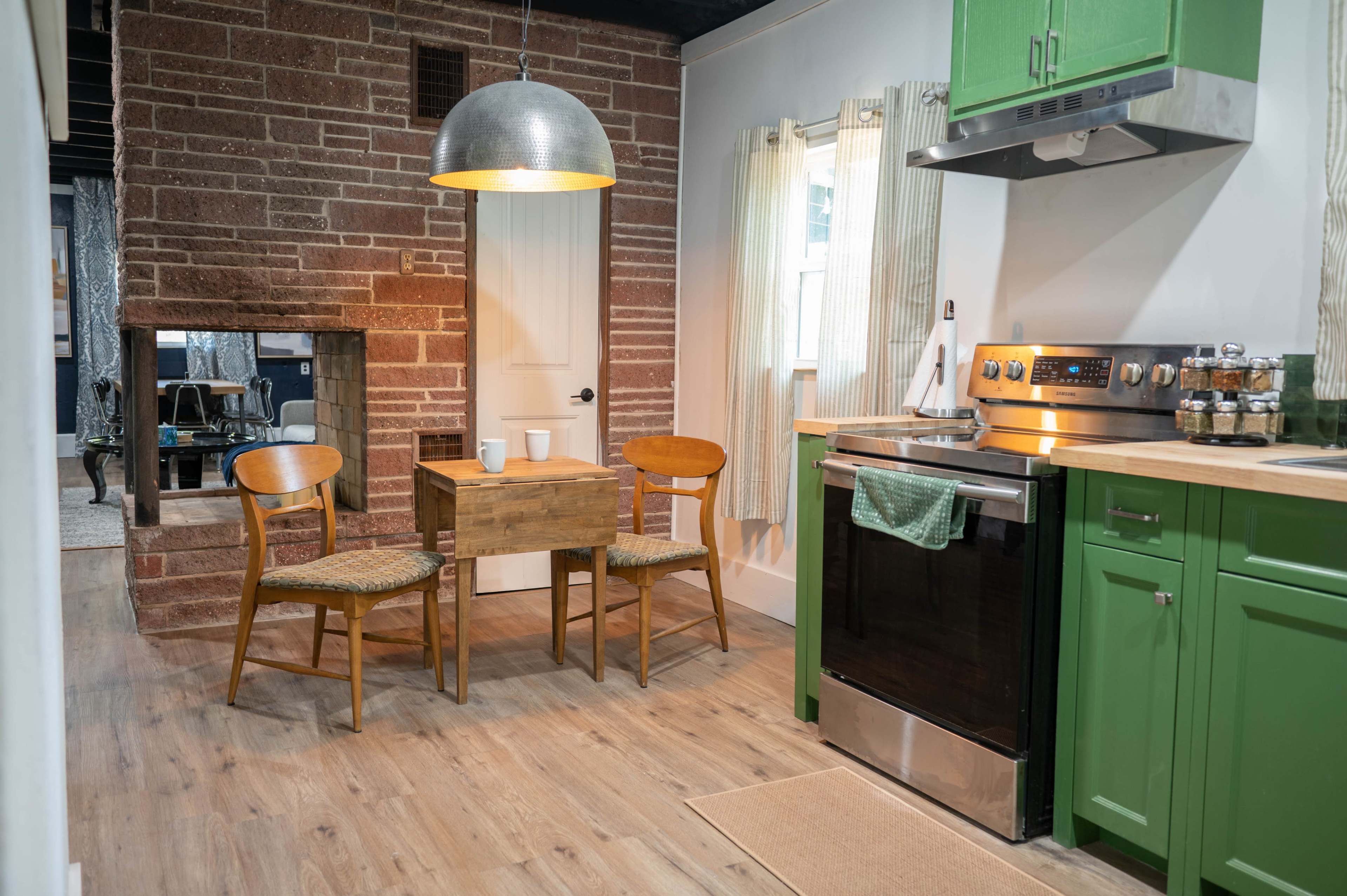 A small kitchen features a wooden table and two chairs, a green cabinetry setup, a stainless steel stove, and a rustic brick wall.