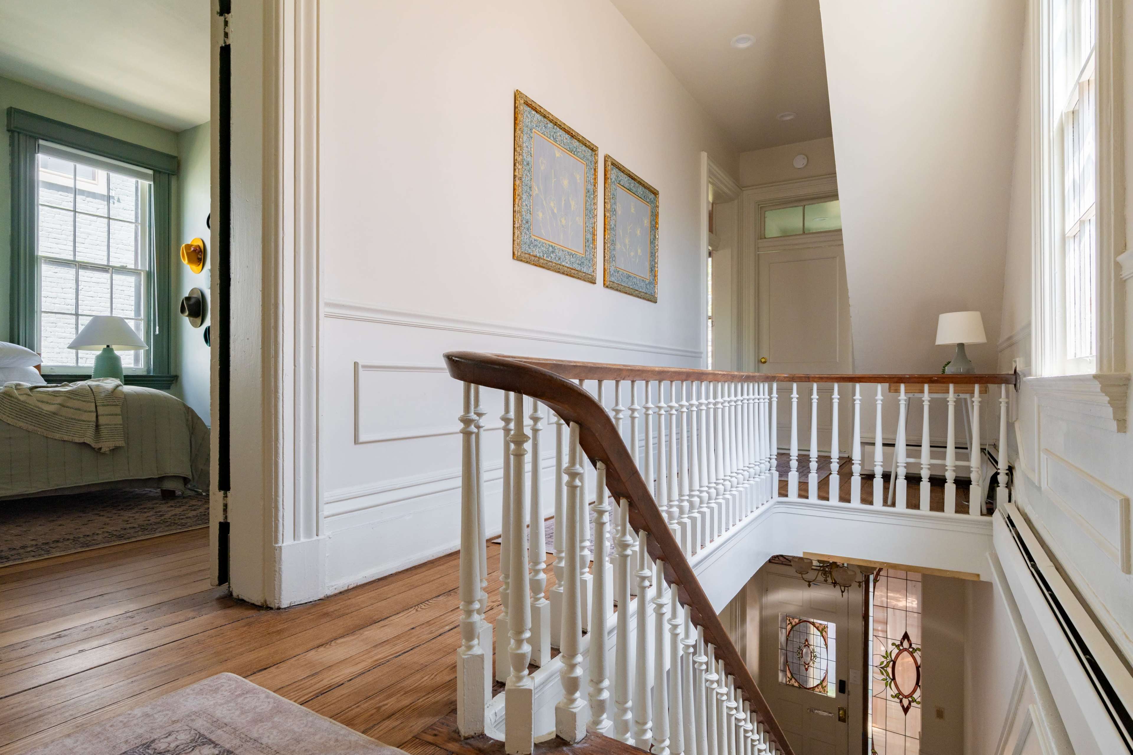 A wooden staircase with a white railing descends into a well-lit hallway, featuring two framed artworks on the wall and a glimpse of a bedroom through an open door.
