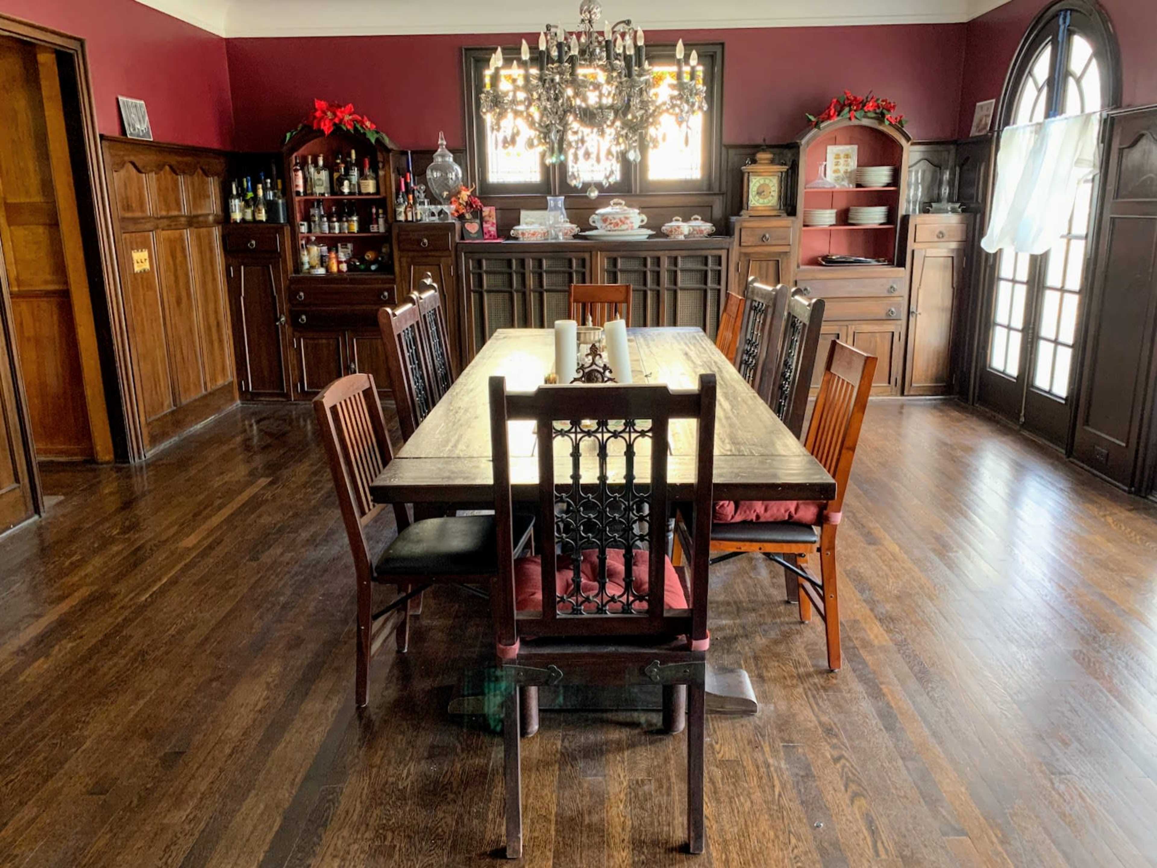 A long wooden dining table surrounded by chairs is positioned in a room with dark red walls and wooden cabinetry, featuring a chandelier overhead and holiday decorations.