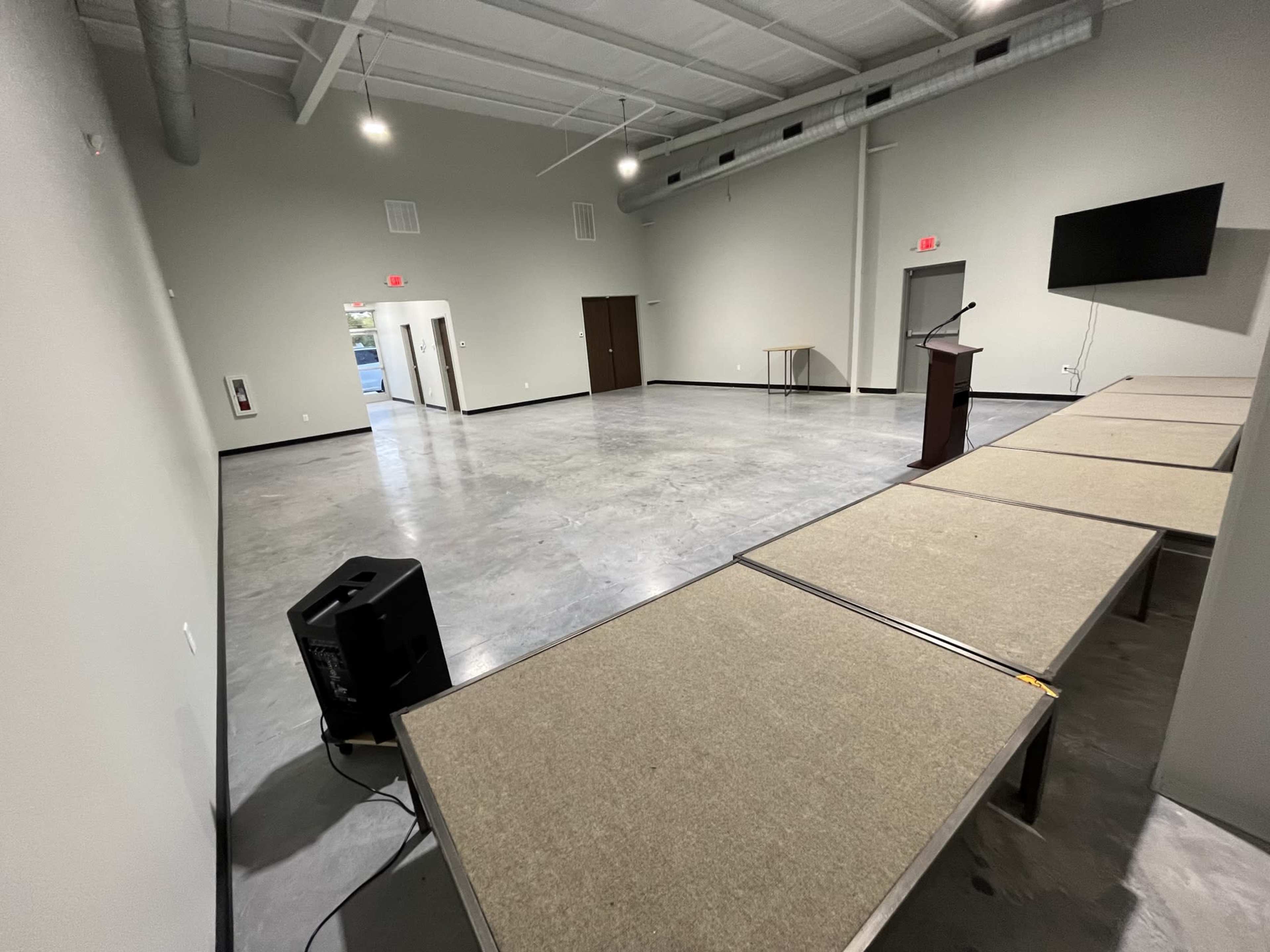 A spacious, empty room with gray floors, tables arranged in a line, and a podium facing a wall-mounted screen.