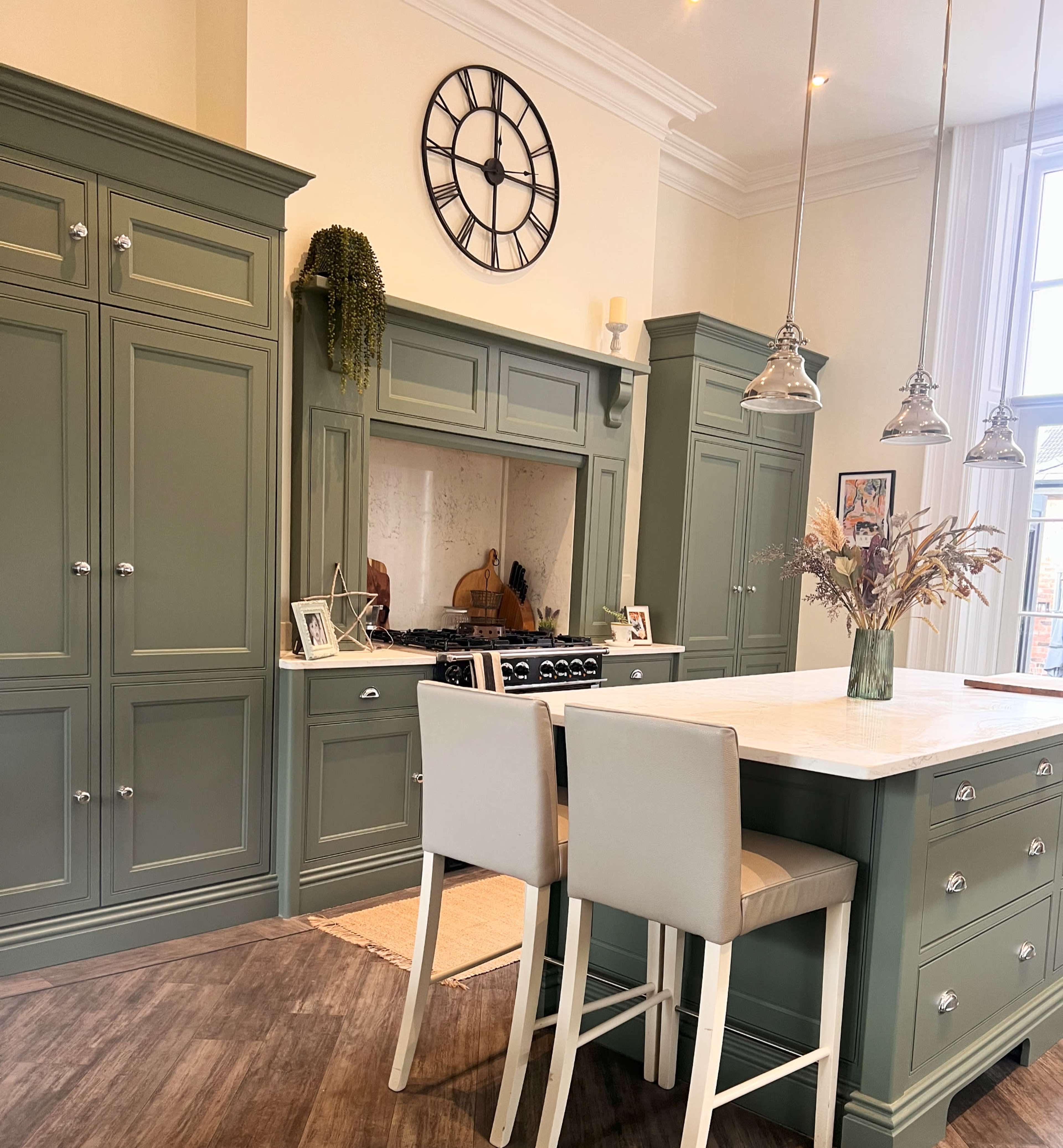 The image depicts a modern kitchen with green cabinetry, a large clock on the wall, a central island with two light-colored stools, and a gas stove framed by a decorative backsplash.