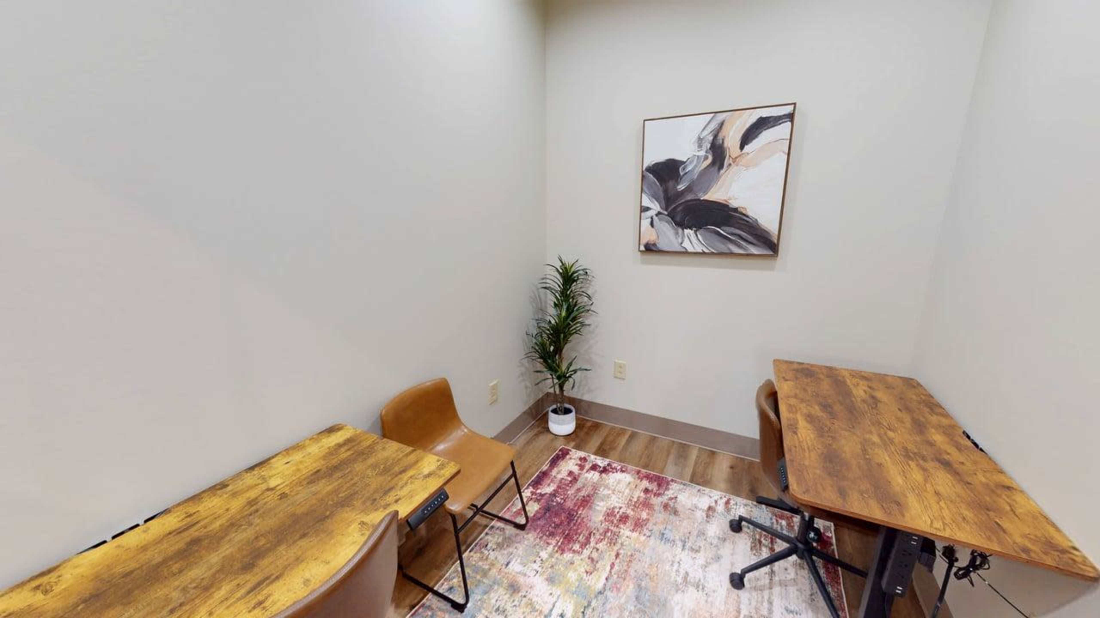 The image shows a small, sparsely furnished room with two wooden tables, two chairs, a potted plant, and a decorative wall art piece.