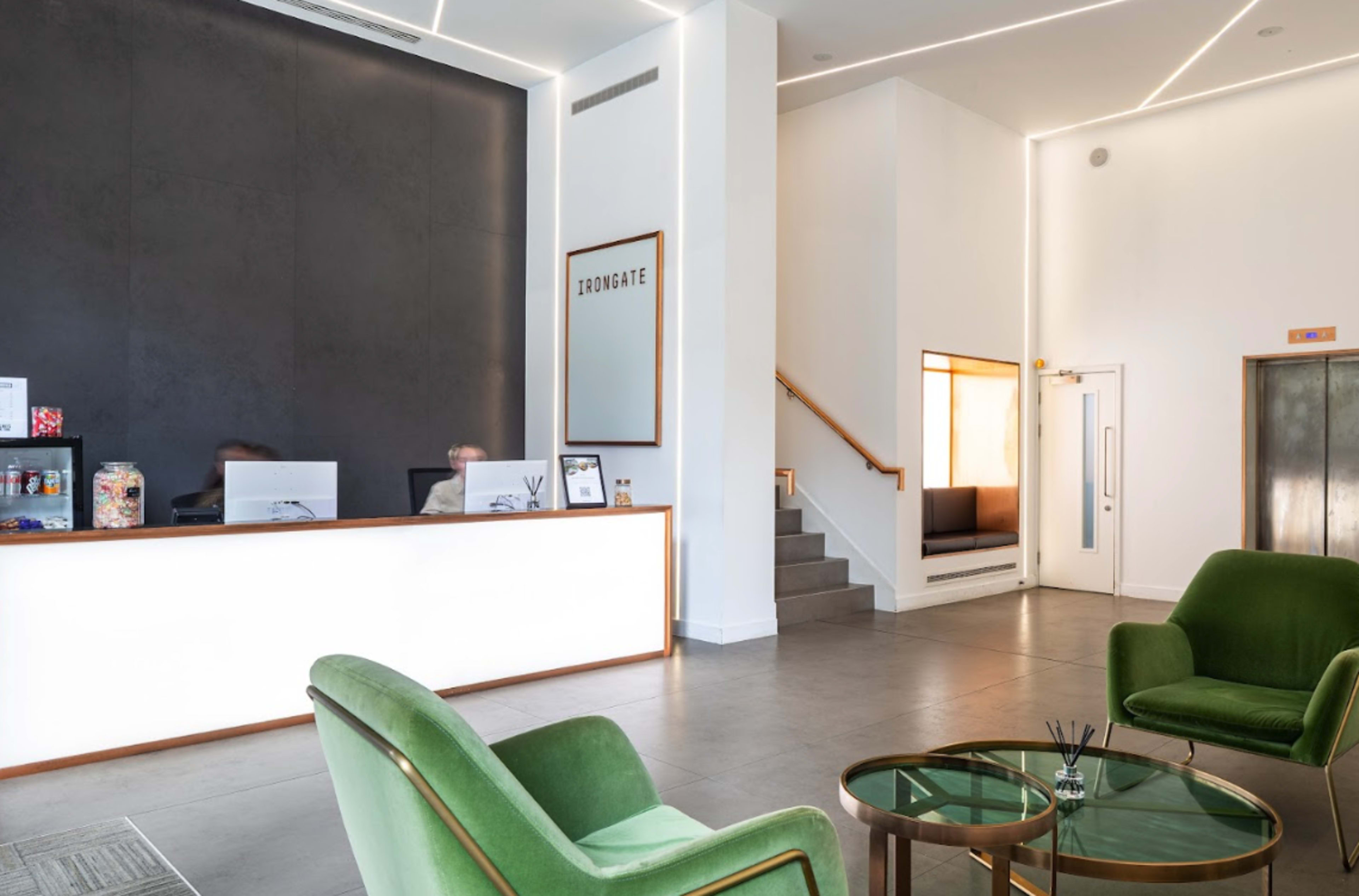 A modern reception area with a sleek front desk, two green chairs, and a staircase leading to a small seating area.