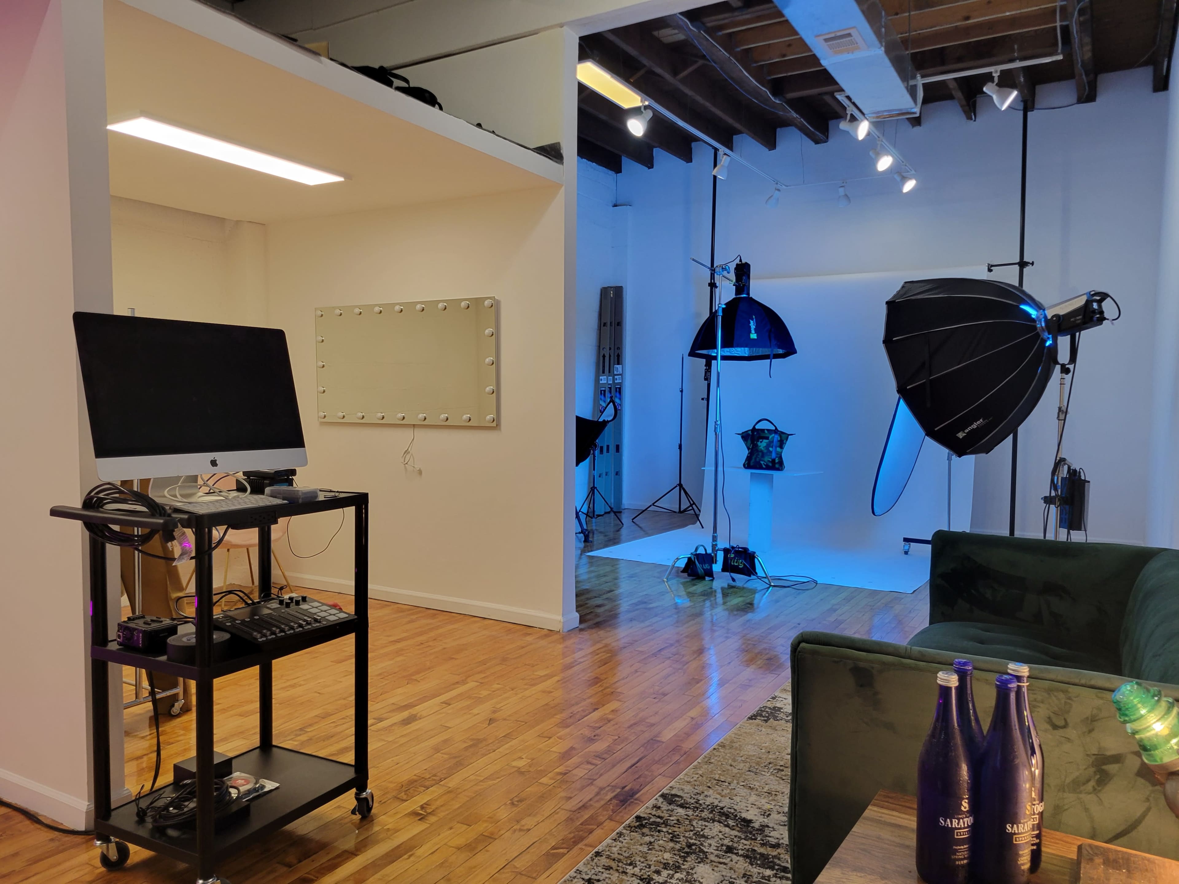 The image shows a well-lit photography studio featuring a large softbox, a backdrop, and a mirrored wall, alongside a seating area with a green couch and a cart with a computer and equipment.