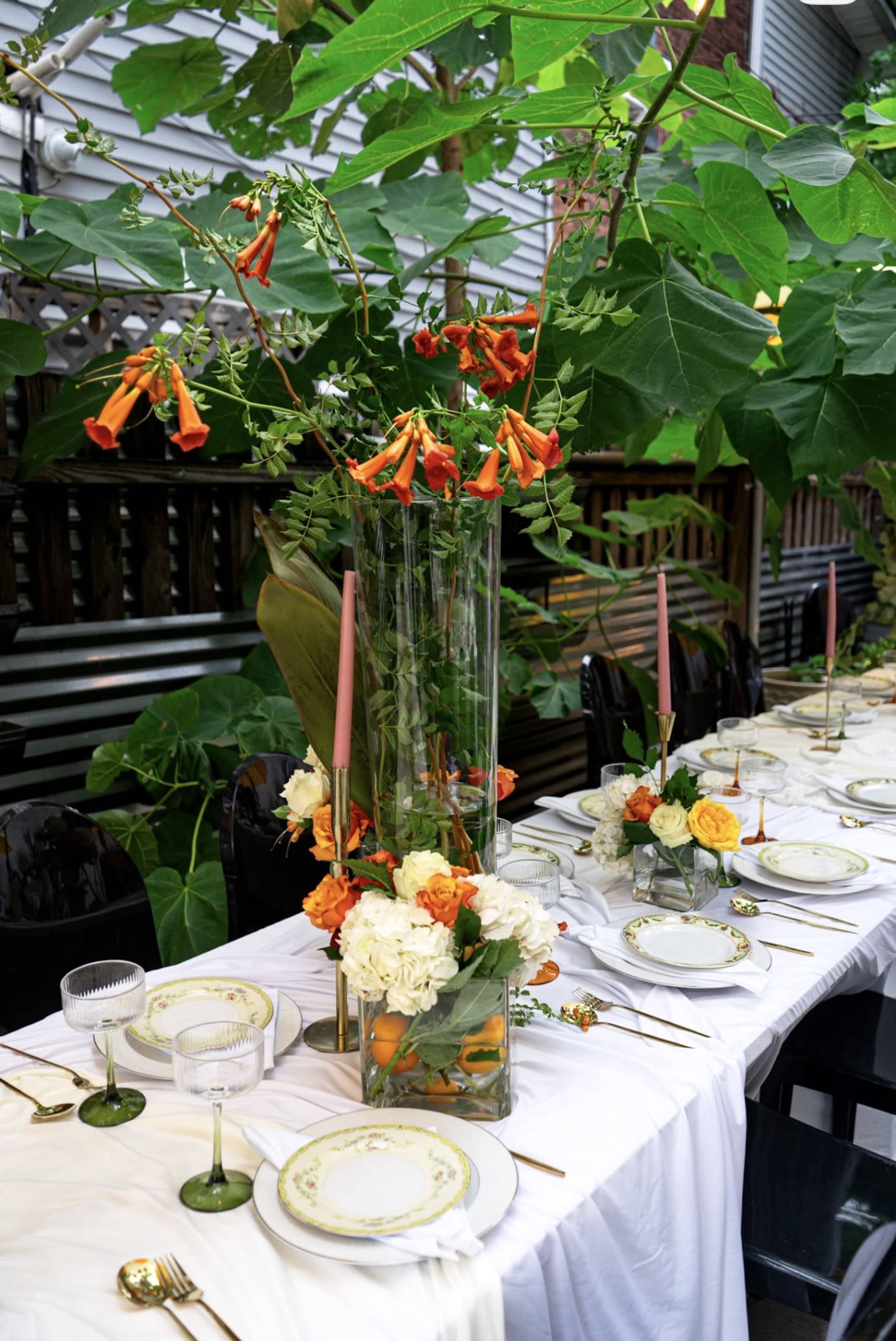 A long dining table is set outdoors with white tablecloths, decorative plates, and a large floral centerpiece featuring orange trumpet flowers and greenery.