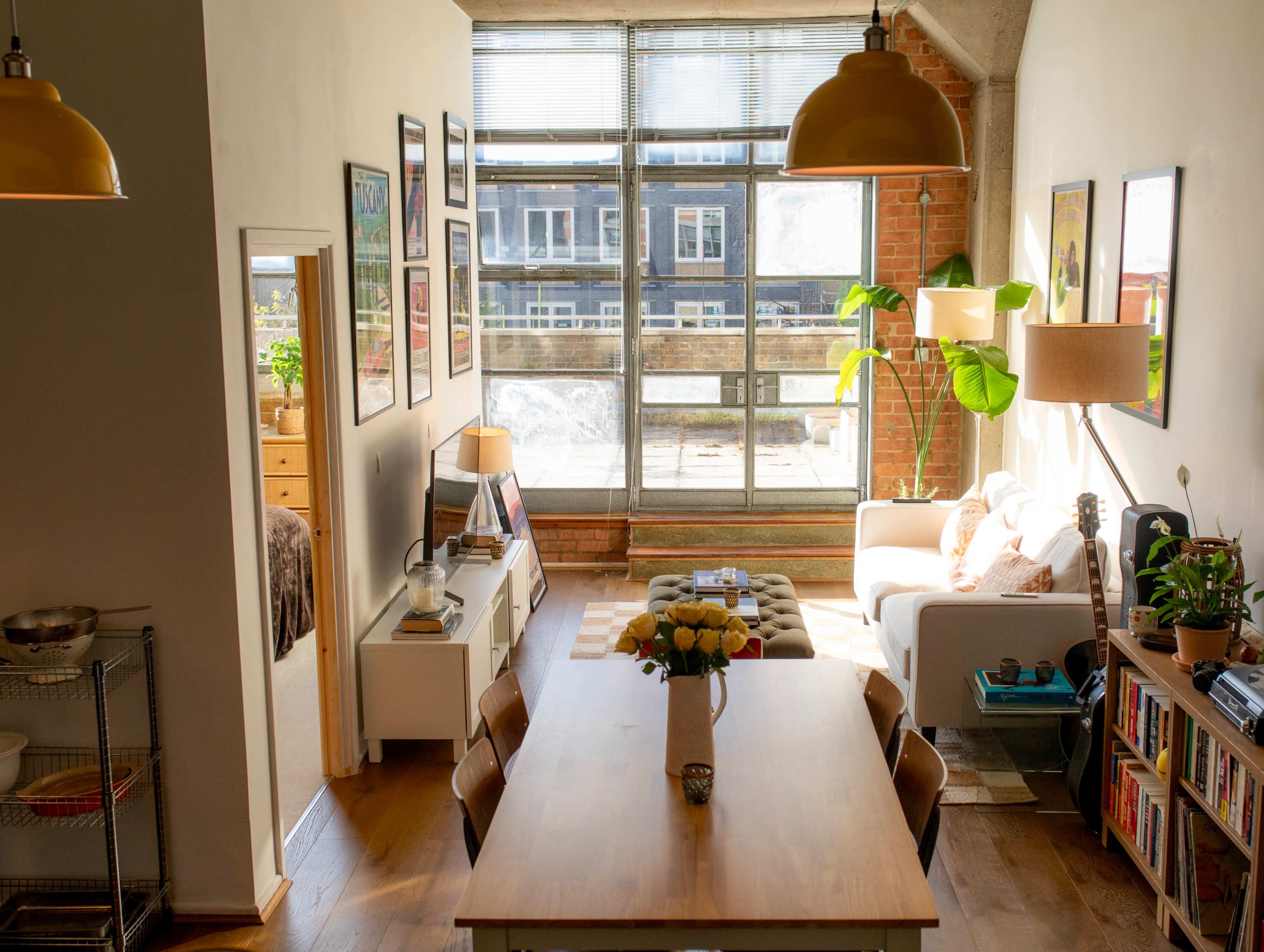 The image shows a well-lit living space featuring a dining table, a sofa near large windows, and decorative plants along with various artworks on the walls.