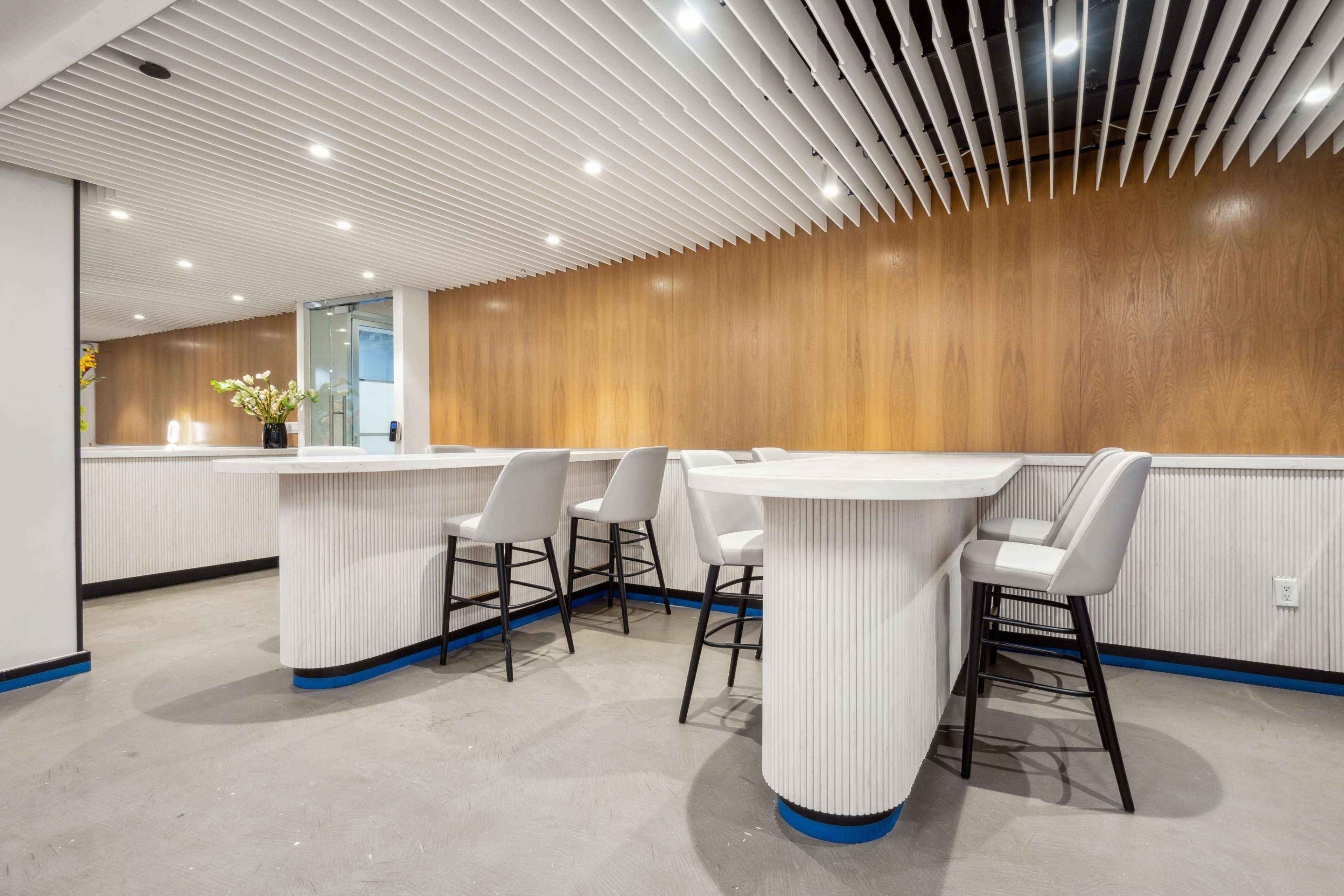 The image shows a modern, minimalist bar area with white countertops, light-colored wood paneling, and high stools.
