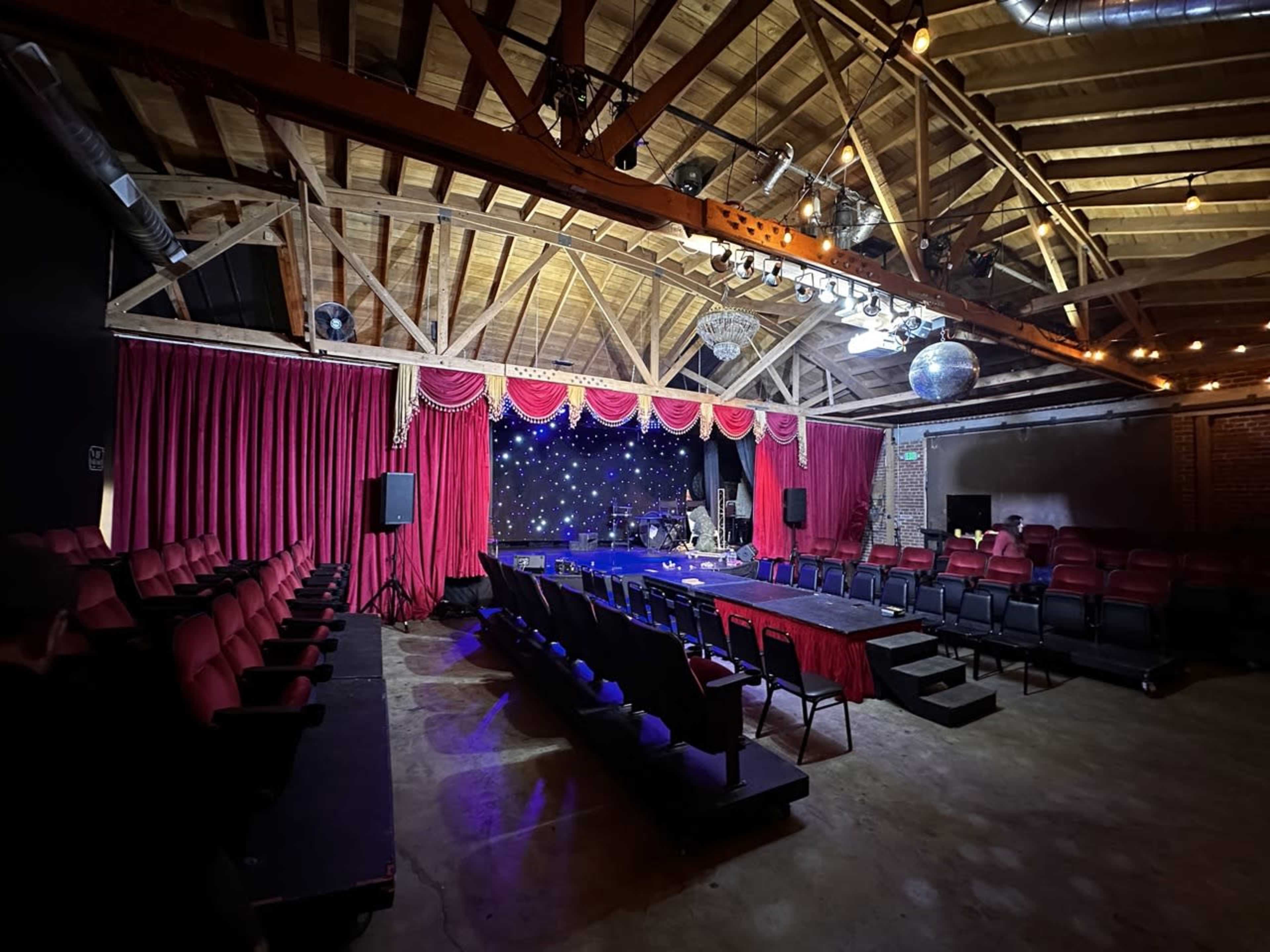 An empty theater with red velvet curtains, a stage adorned with twinkling lights, and rows of red and black seats arranged for an audience.