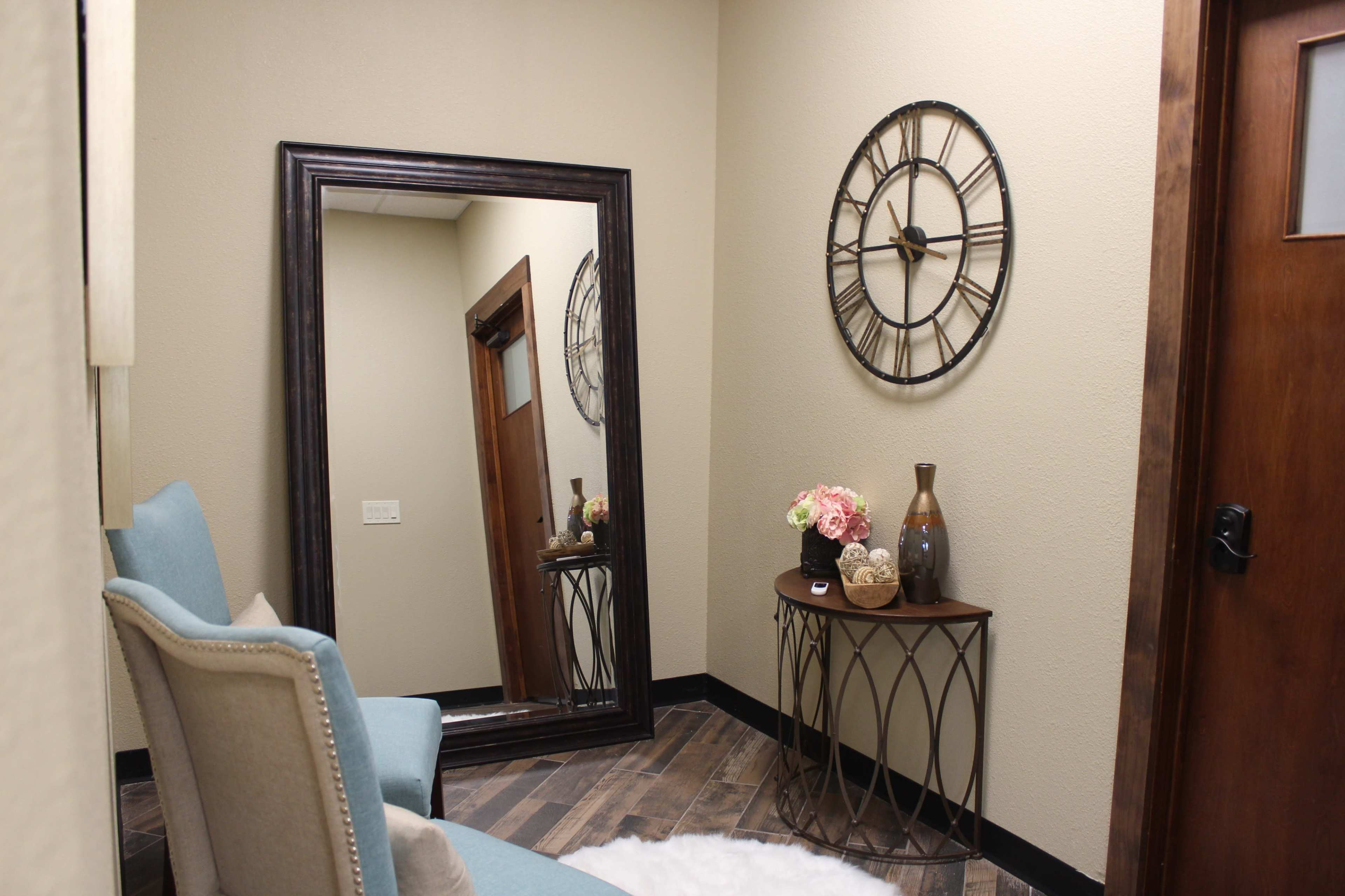 A hallway with a large mirror, a decorative wall clock, a small table with a vase and flowers, and a pair of upholstered chairs against a neutral wall.