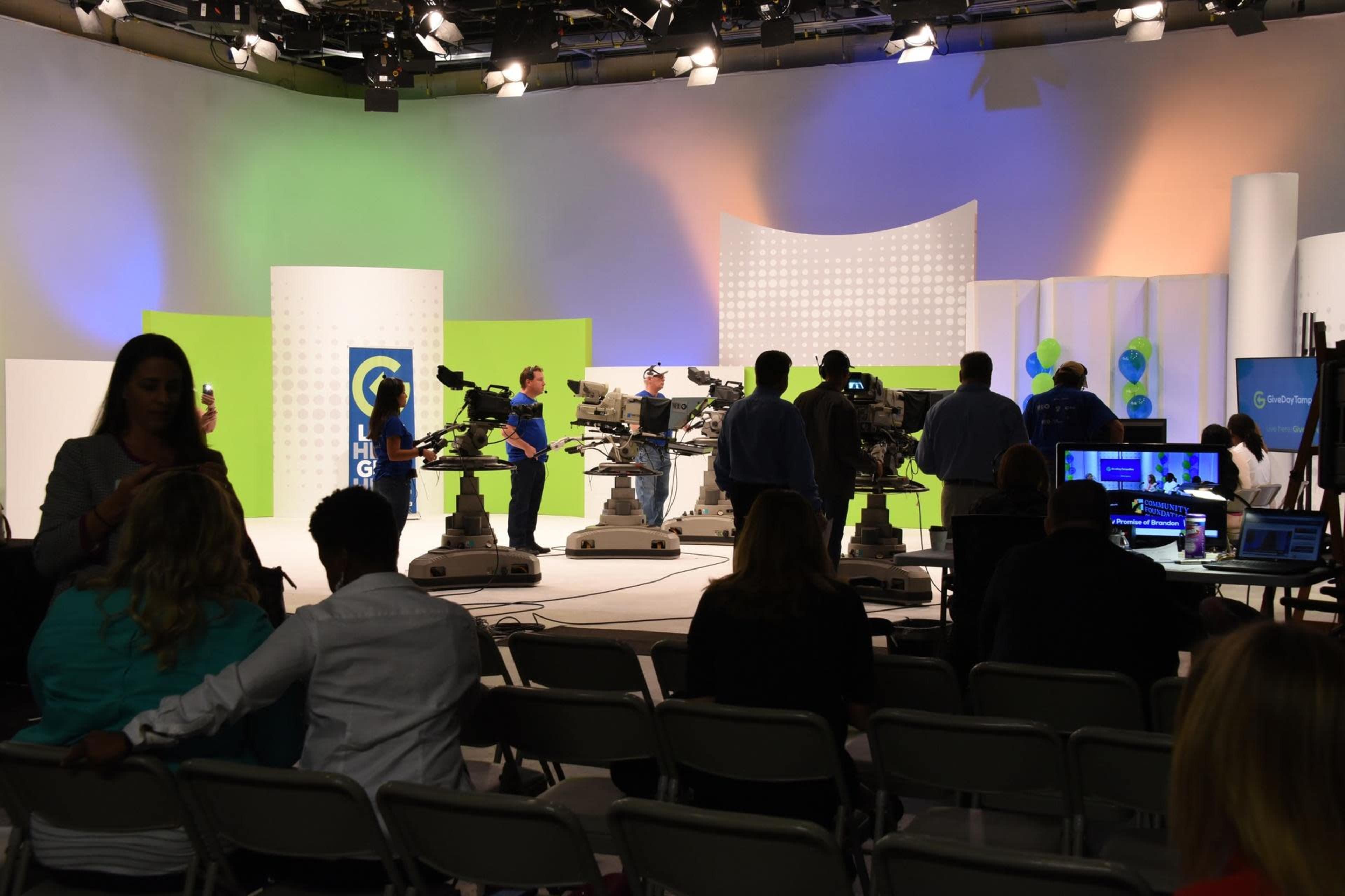 A television studio is set up for a broadcast, with multiple cameras positioned on dollies and crew members preparing the scene, while an audience sits in rows of chairs.