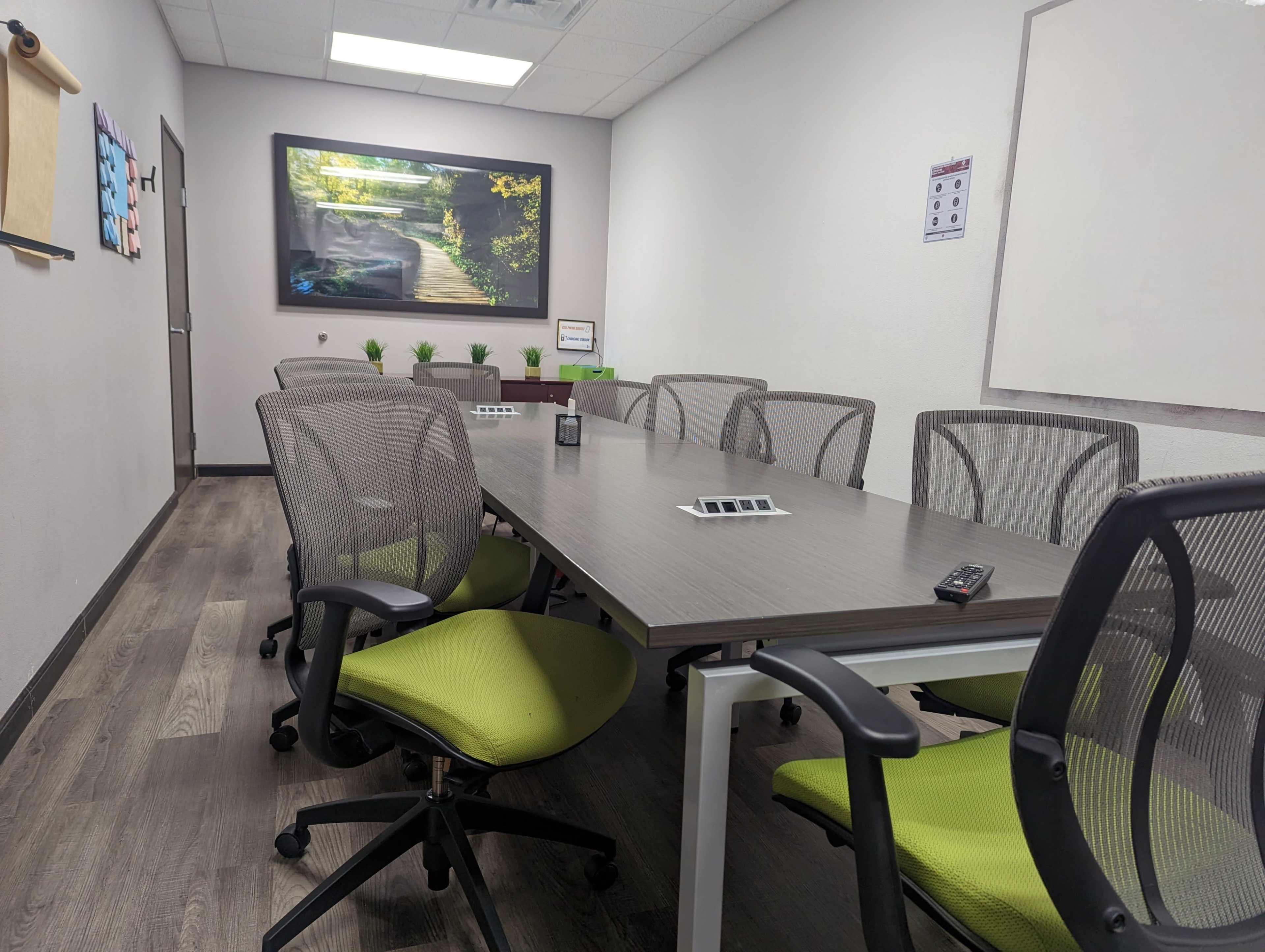 The image shows a modern conference room with a long table surrounded by ergonomic chairs and a large screen displaying a nature scene on the wall.