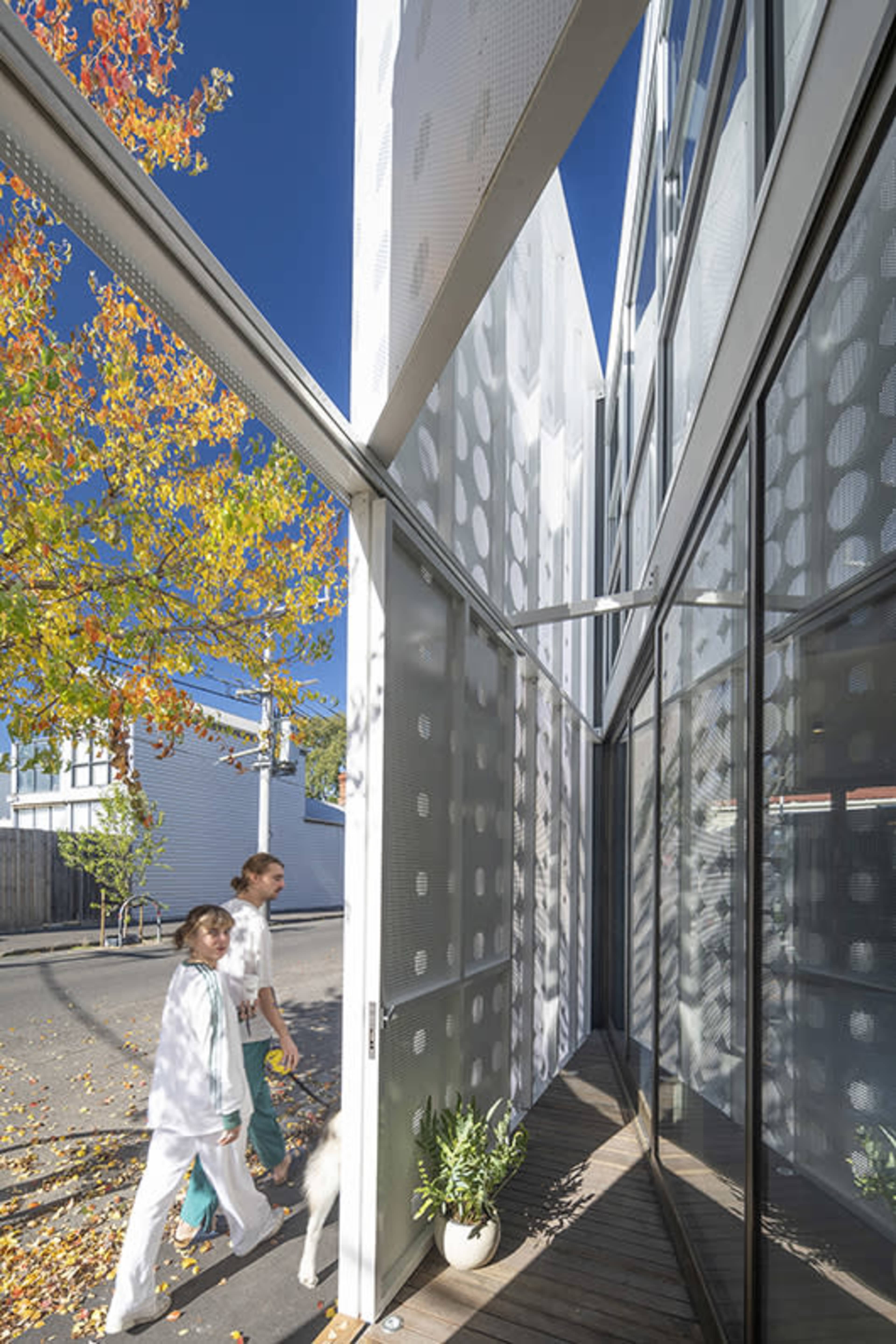 Two people walk by a modern building with large glass windows and patterned metal panels, surrounded by autumn foliage.