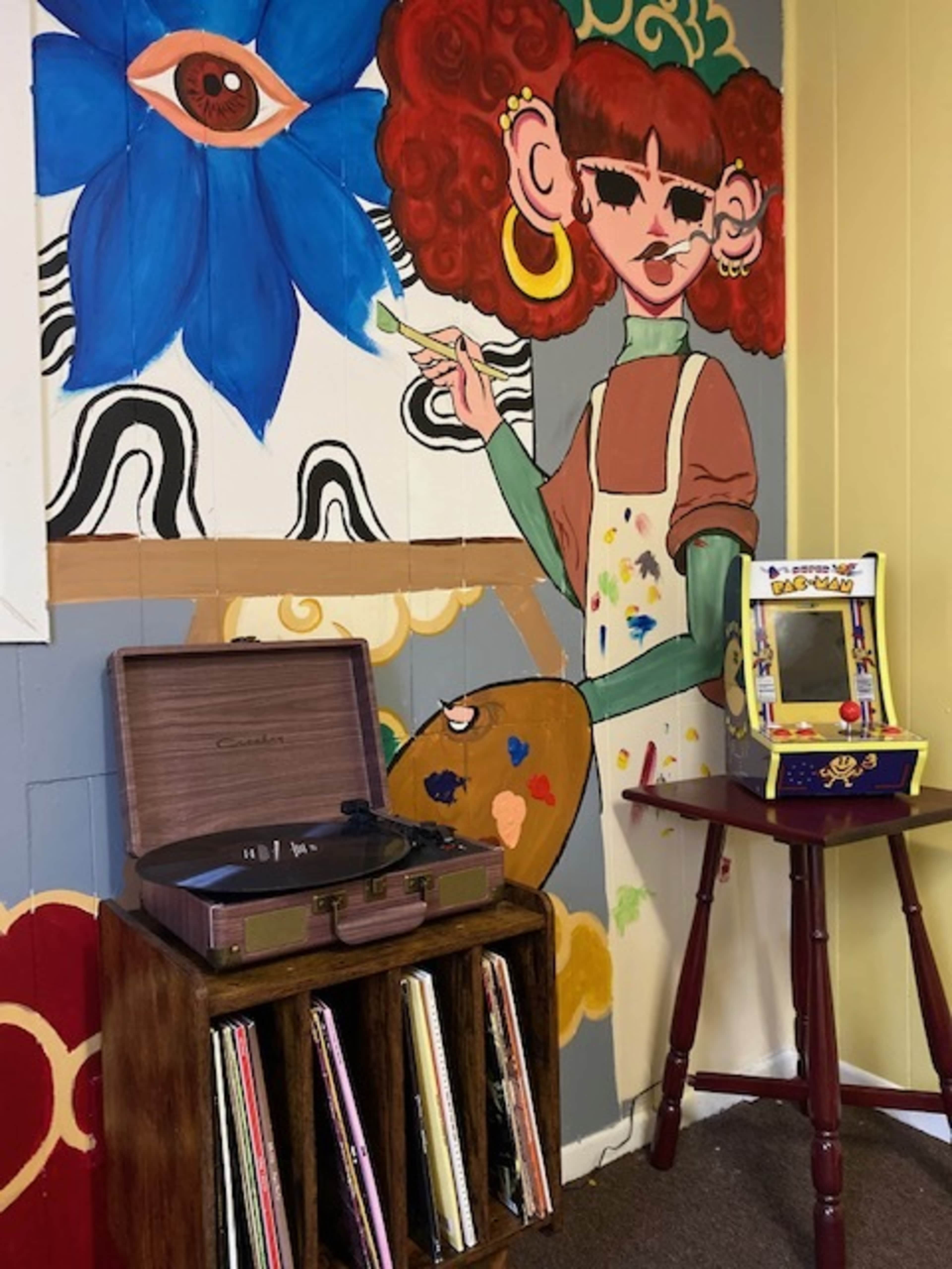 A colorful mural featuring a girl with curly hair and an eye is painted on the wall next to a vintage record player and a small mirrored console table.