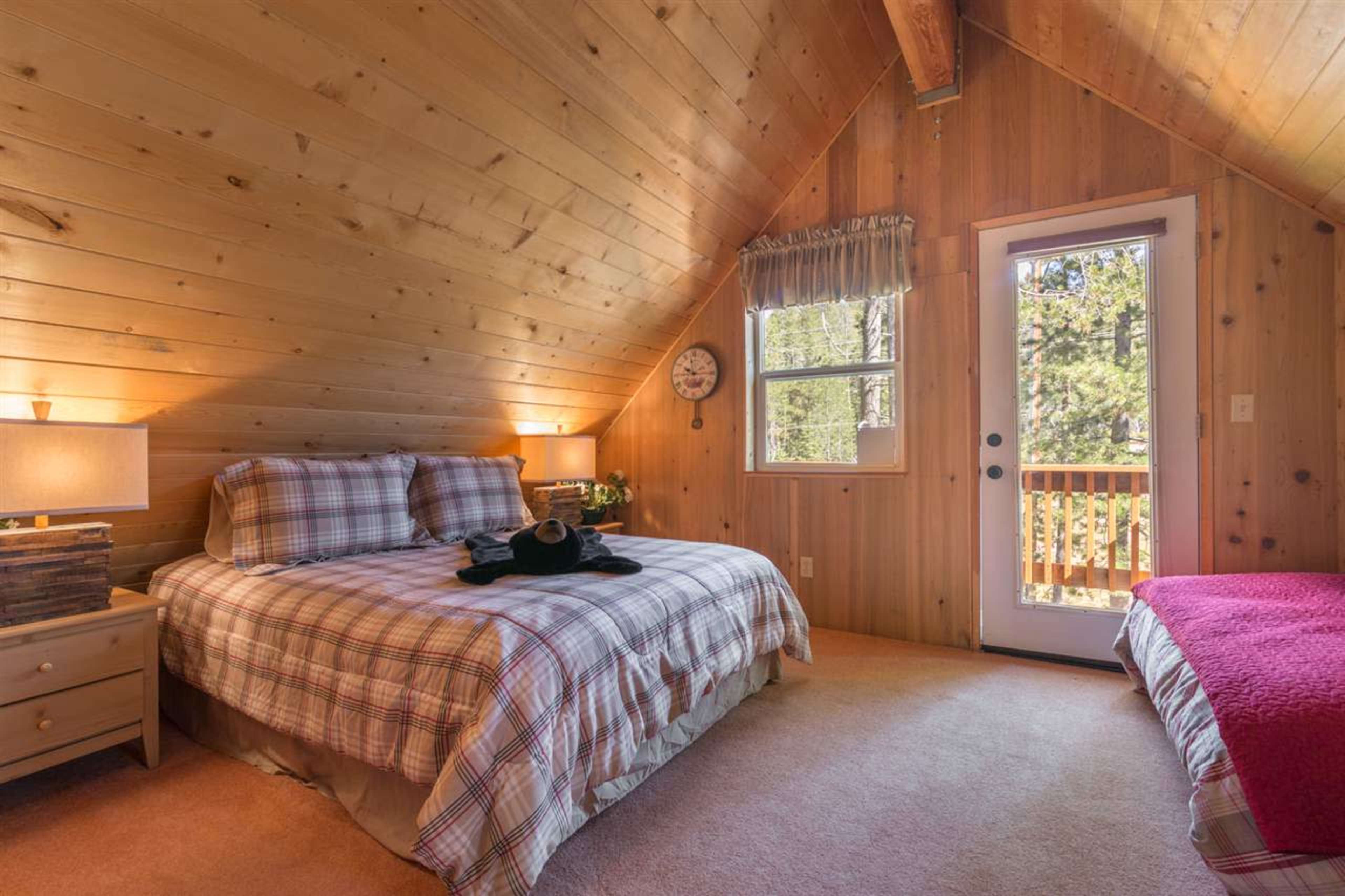 The image shows a cozy bedroom with wooden walls, a queen-sized bed covered in a plaid comforter, and a single red bed near a window with a door leading to a balcony.