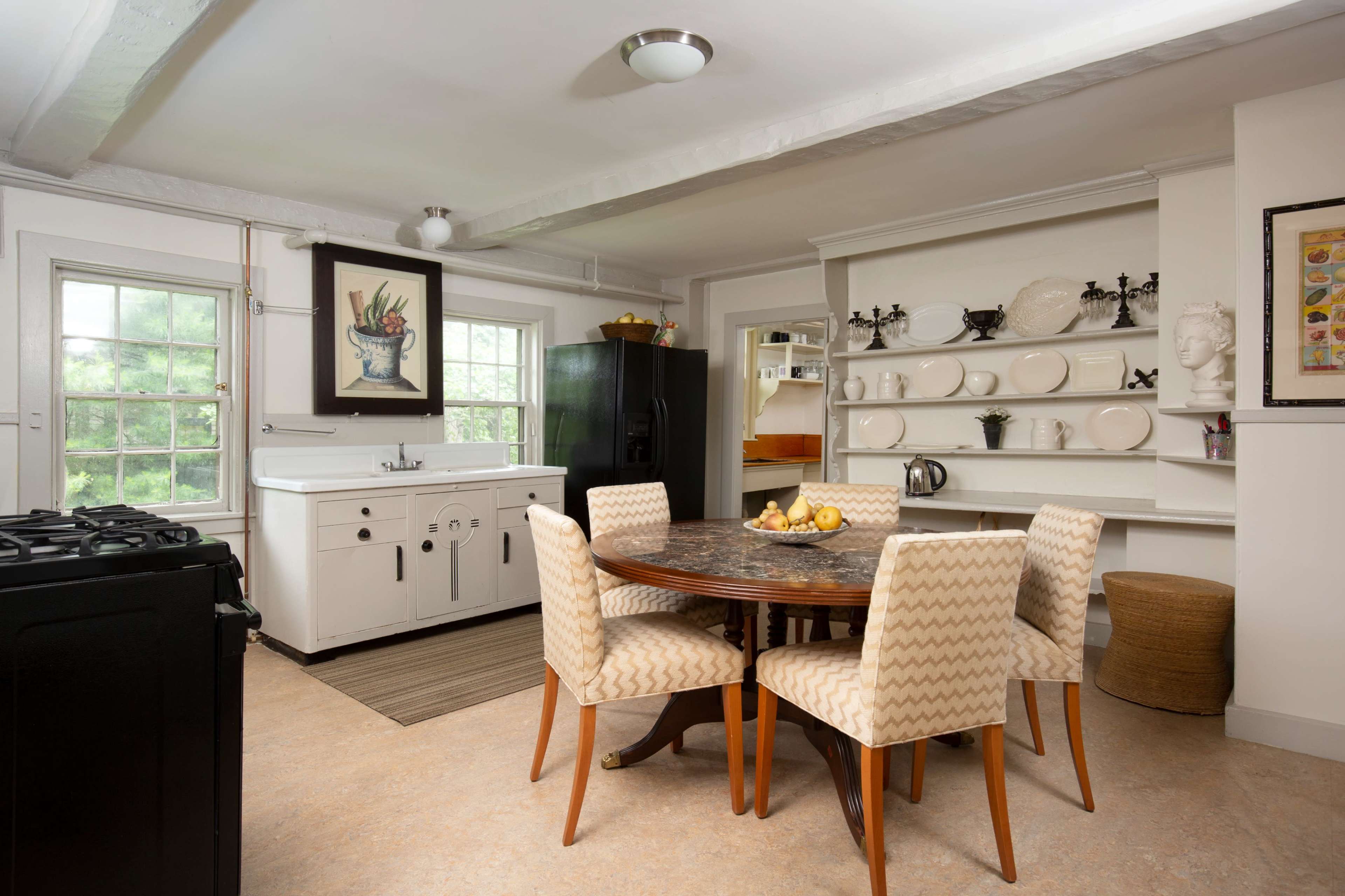 A kitchen with a round wooden table surrounded by four upholstered chairs, a black gas stove, a white sink and countertop, and shelves displaying plates and a kettle against a light-colored wall.