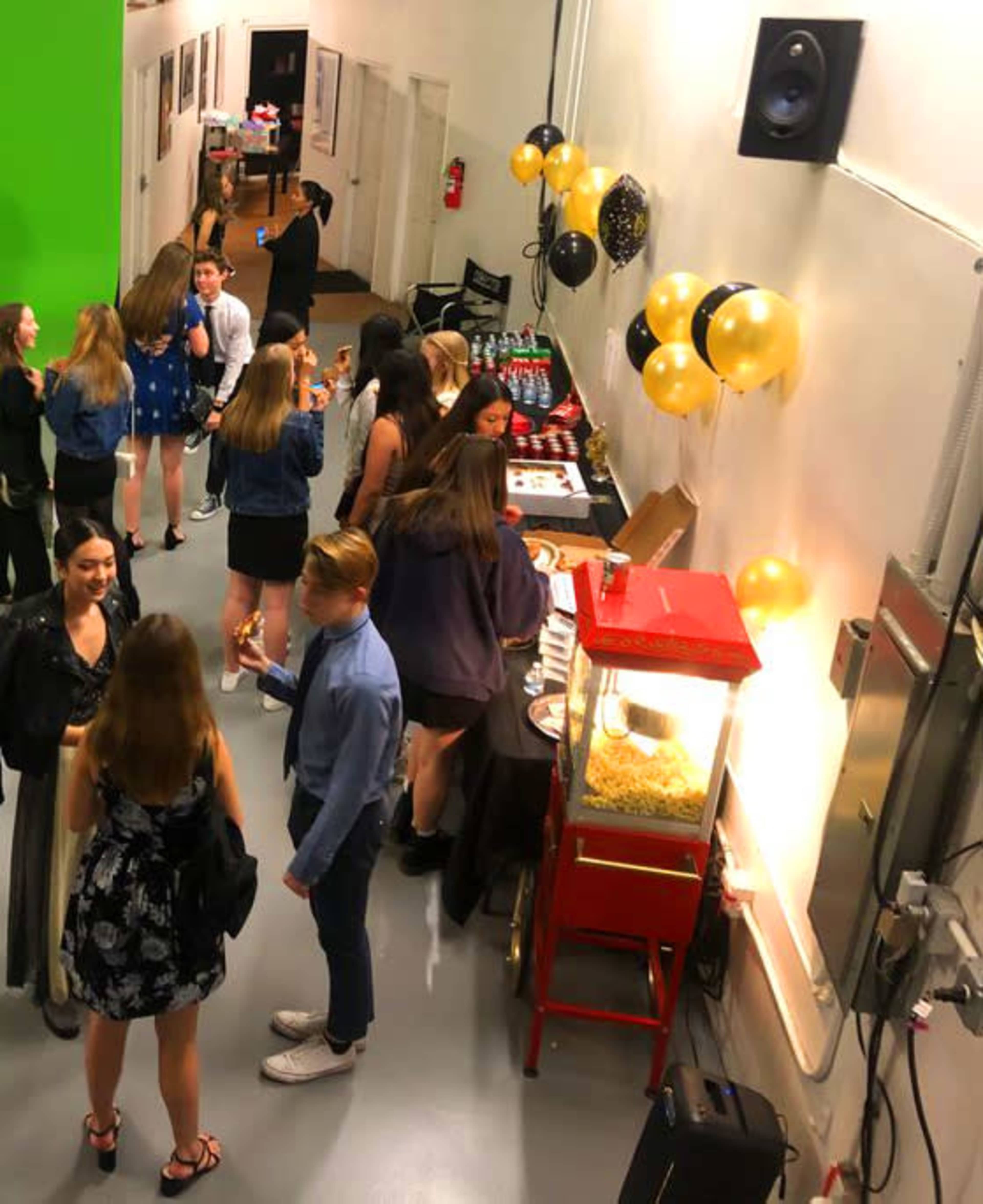 A group of people is gathered in a brightly lit indoor space with a food table and a popcorn machine, decorated with black and gold balloons.